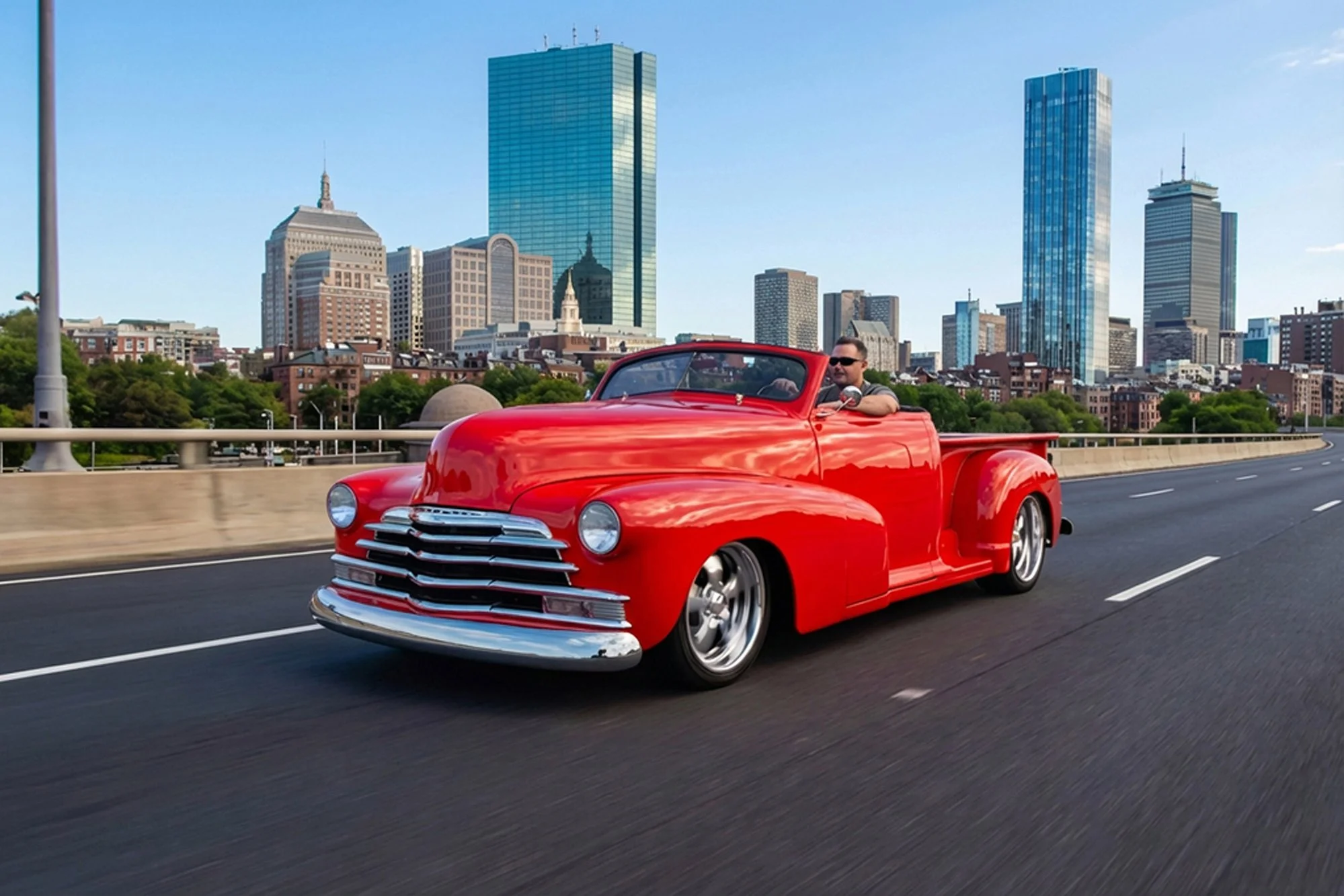 A man driving a vintage red convertible car on a city highway with tall skyscrapers in the background on a clear day.