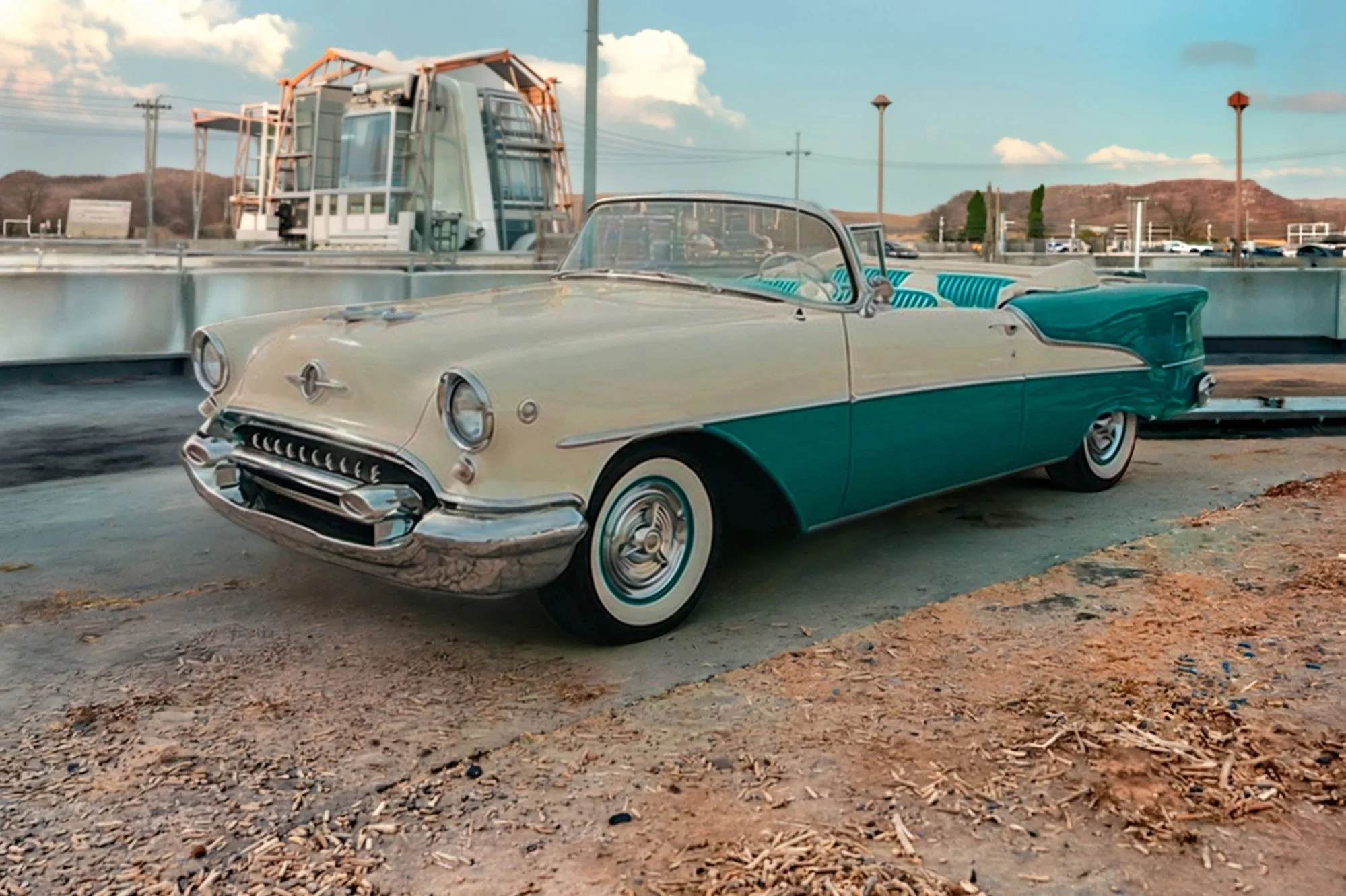 A vintage two-tone teal and cream convertible car parked on a dirt lot with a modern building and power lines in the background under a partly cloudy sky.
