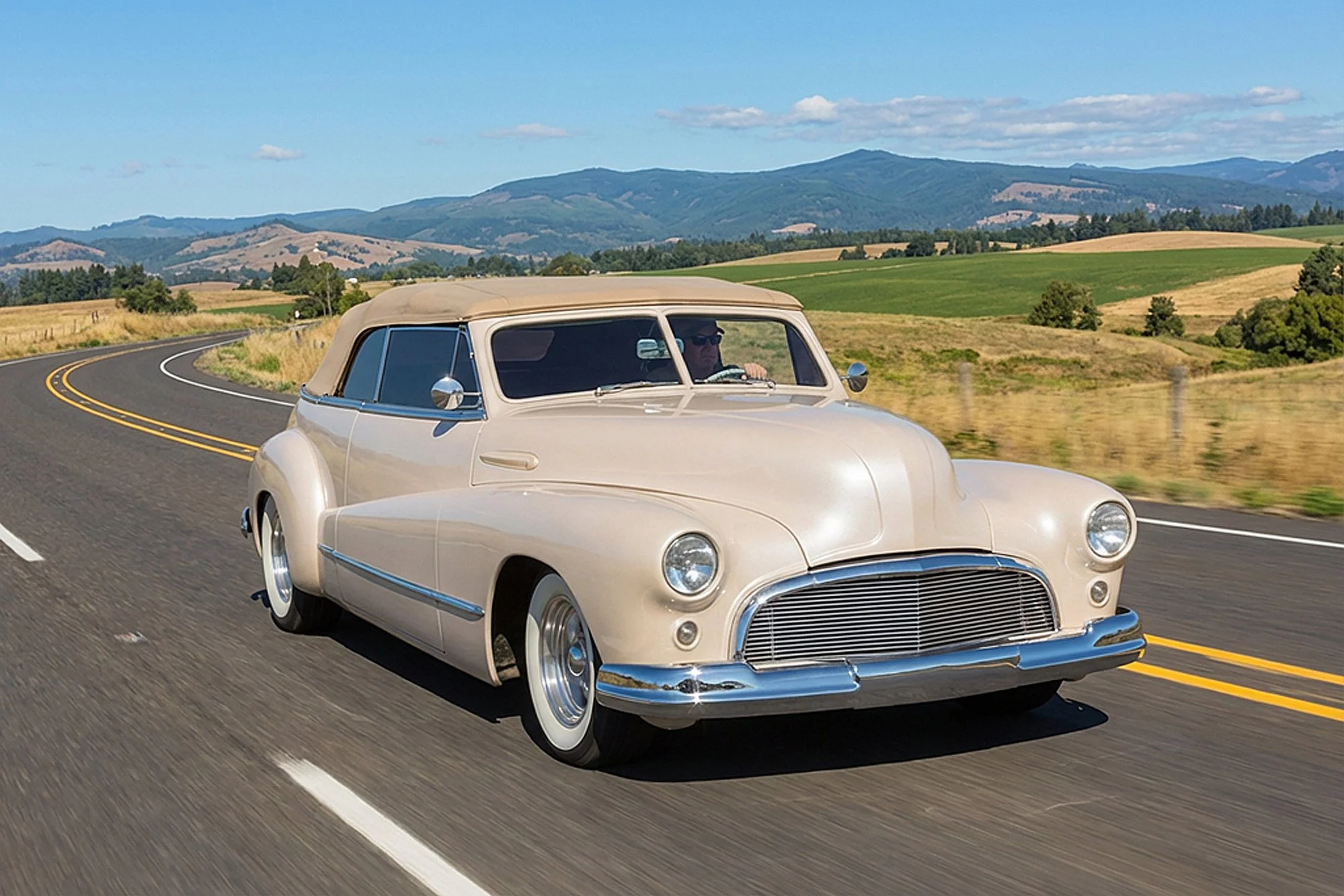 A vintage cream-colored convertible car with a beige soft top driving on a winding two-lane road through a rural landscape with green and brown fields and distant mountains under a blue sky with scattered clouds.
