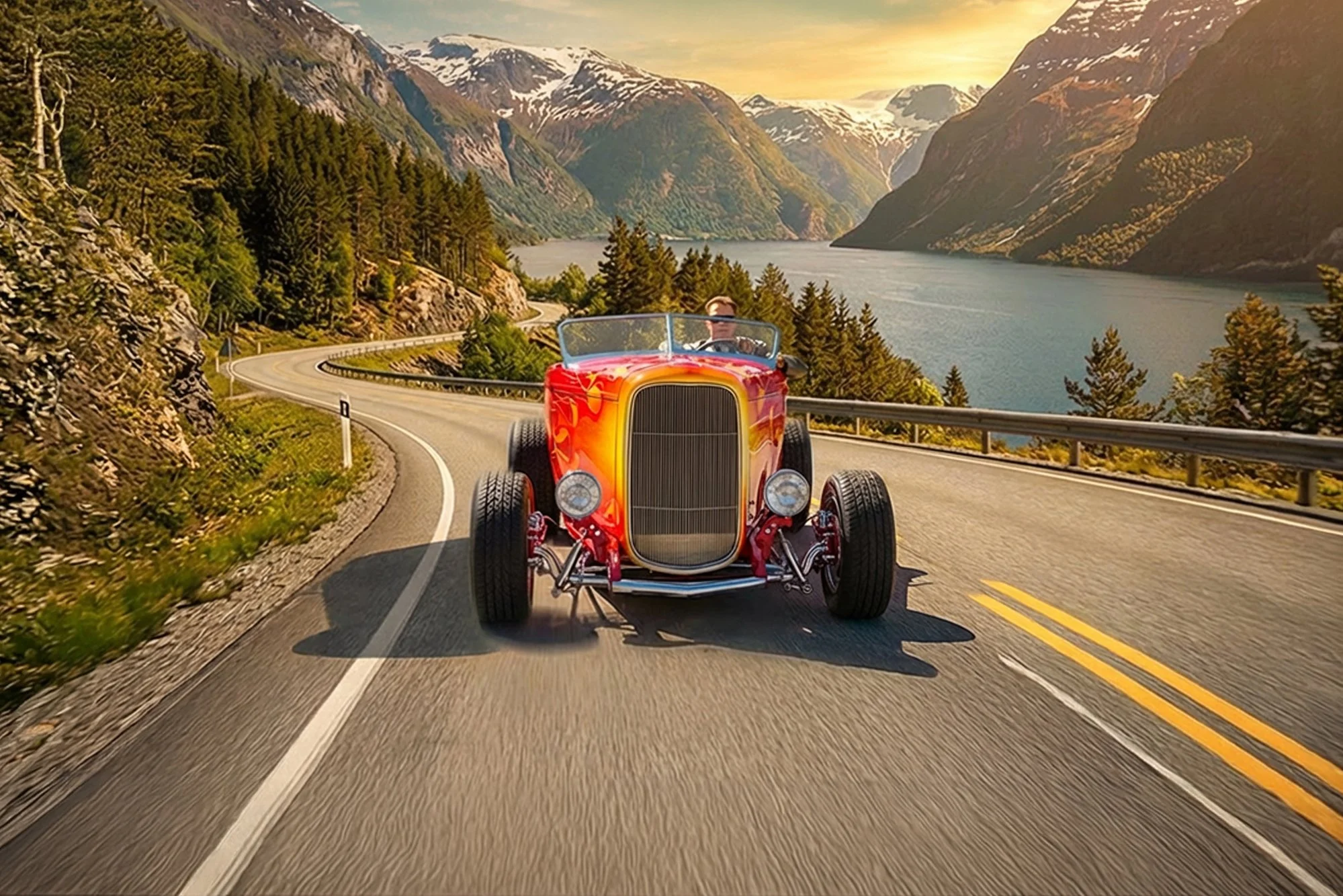 A person driving a vintage red convertible car on a winding mountain road surrounded by trees and overlooking a lake and snow-capped mountains at sunset.