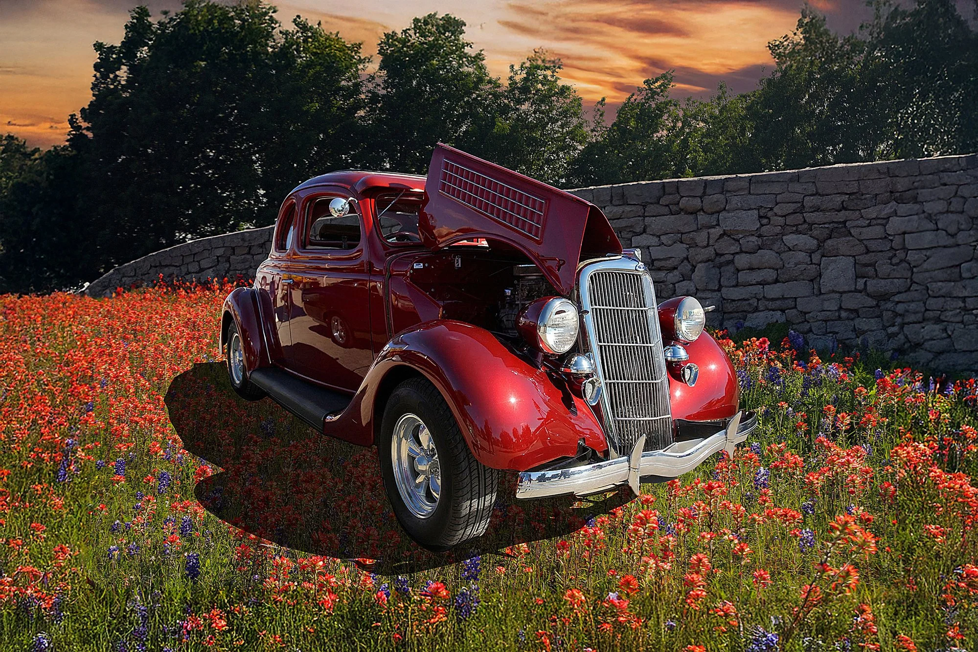 A vintage red car with its hood open, sitting in a colorful flower field at sunset with trees and a stone wall in the background.