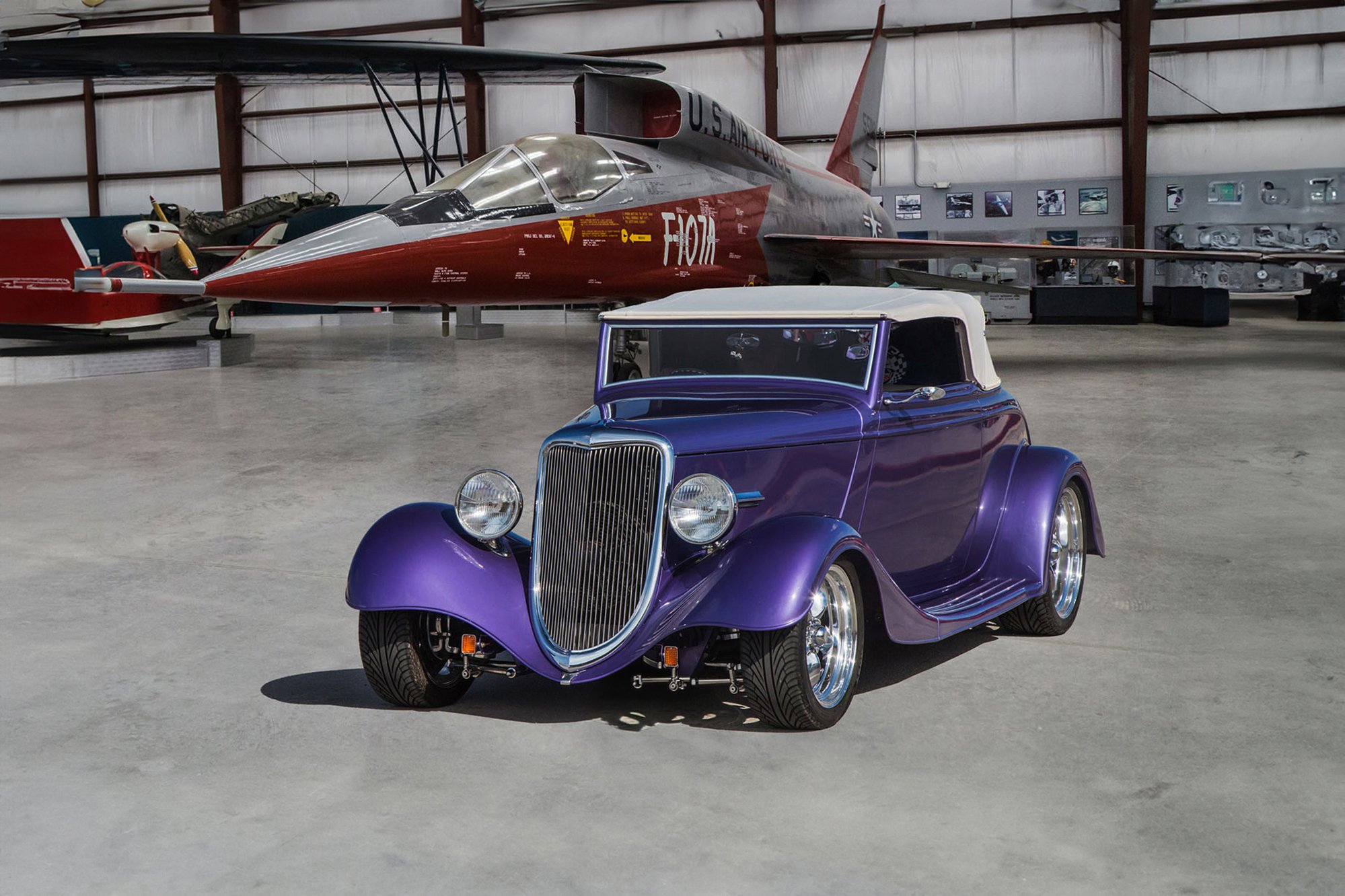 A vintage purple convertible car parked inside a museum with a large aircraft in the background.
