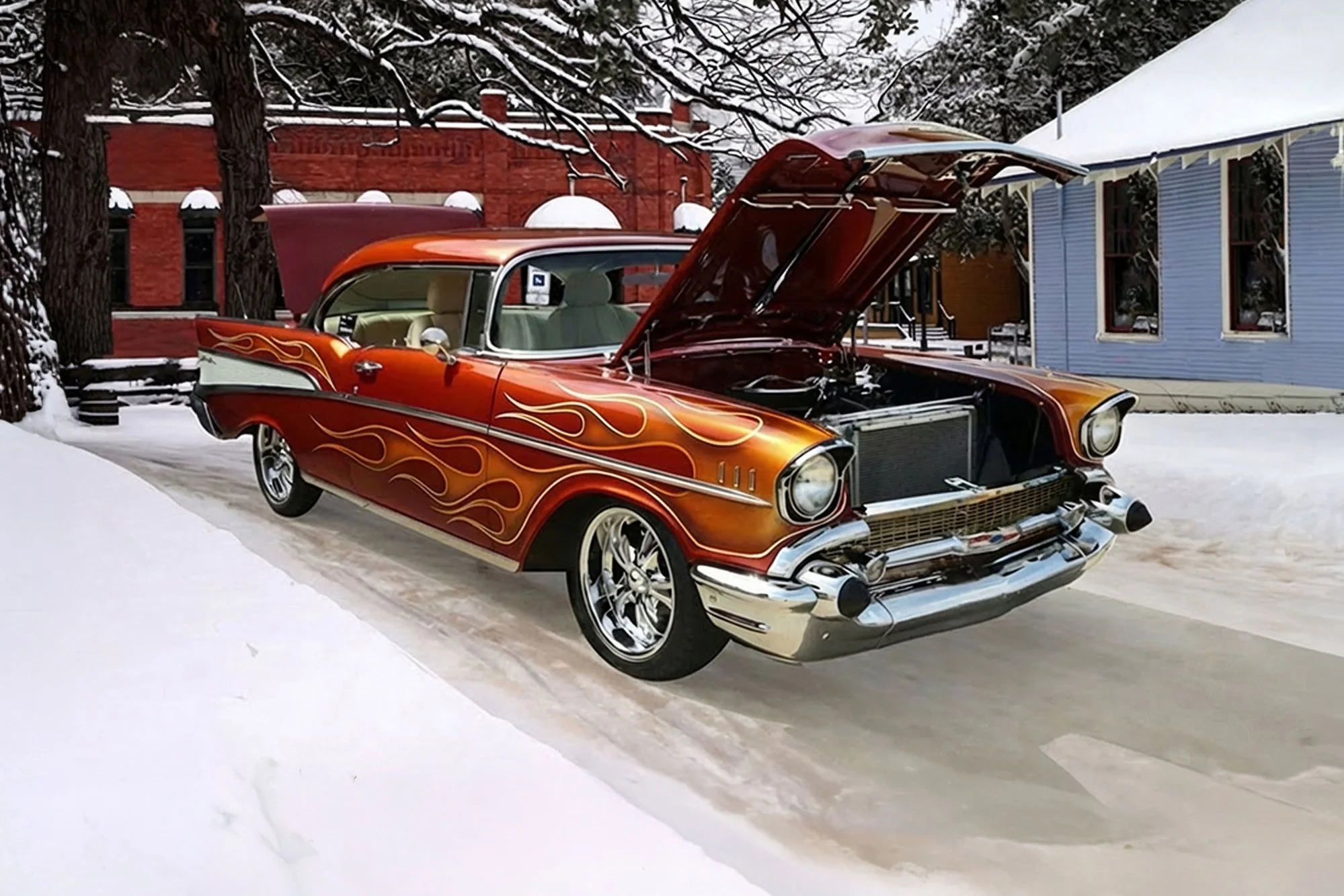 A vintage red and orange car with flame decals has its hood open, parked on snow in front of a blue house and a red building after a snowstorm during daytime.