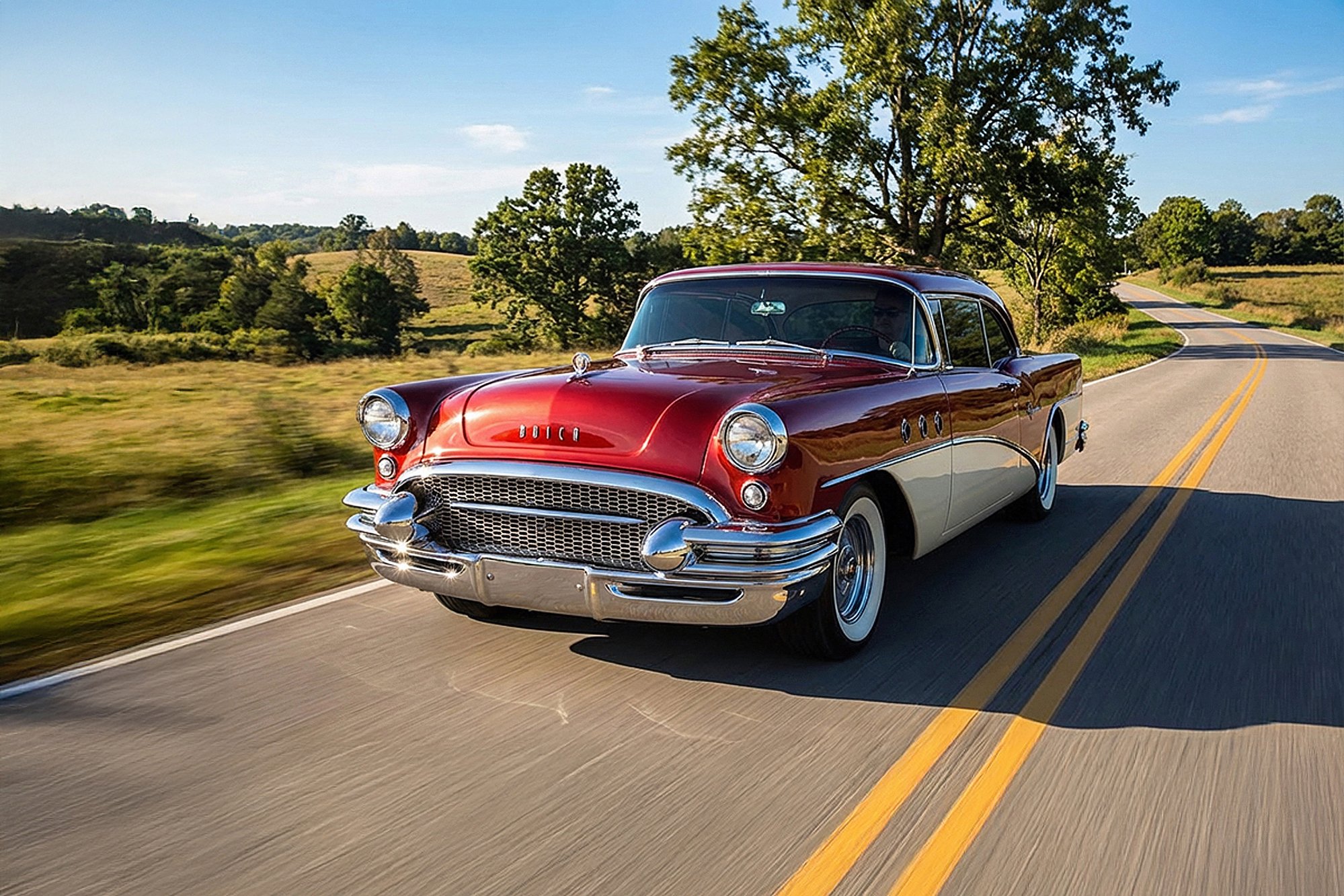 A classic red and white vintage Buick car driving down a winding country road surrounded by green trees and fields under a clear blue sky.