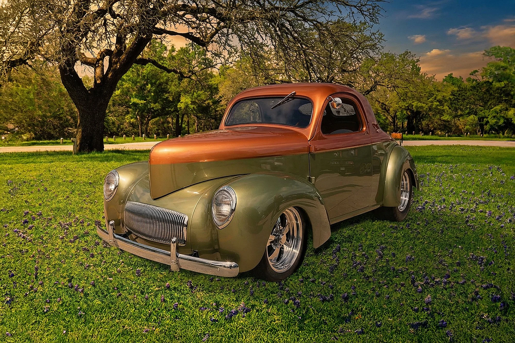A vintage green and brown car parked on a grassy field with purple flowers, under a large tree with sprawling branches, during sunset.