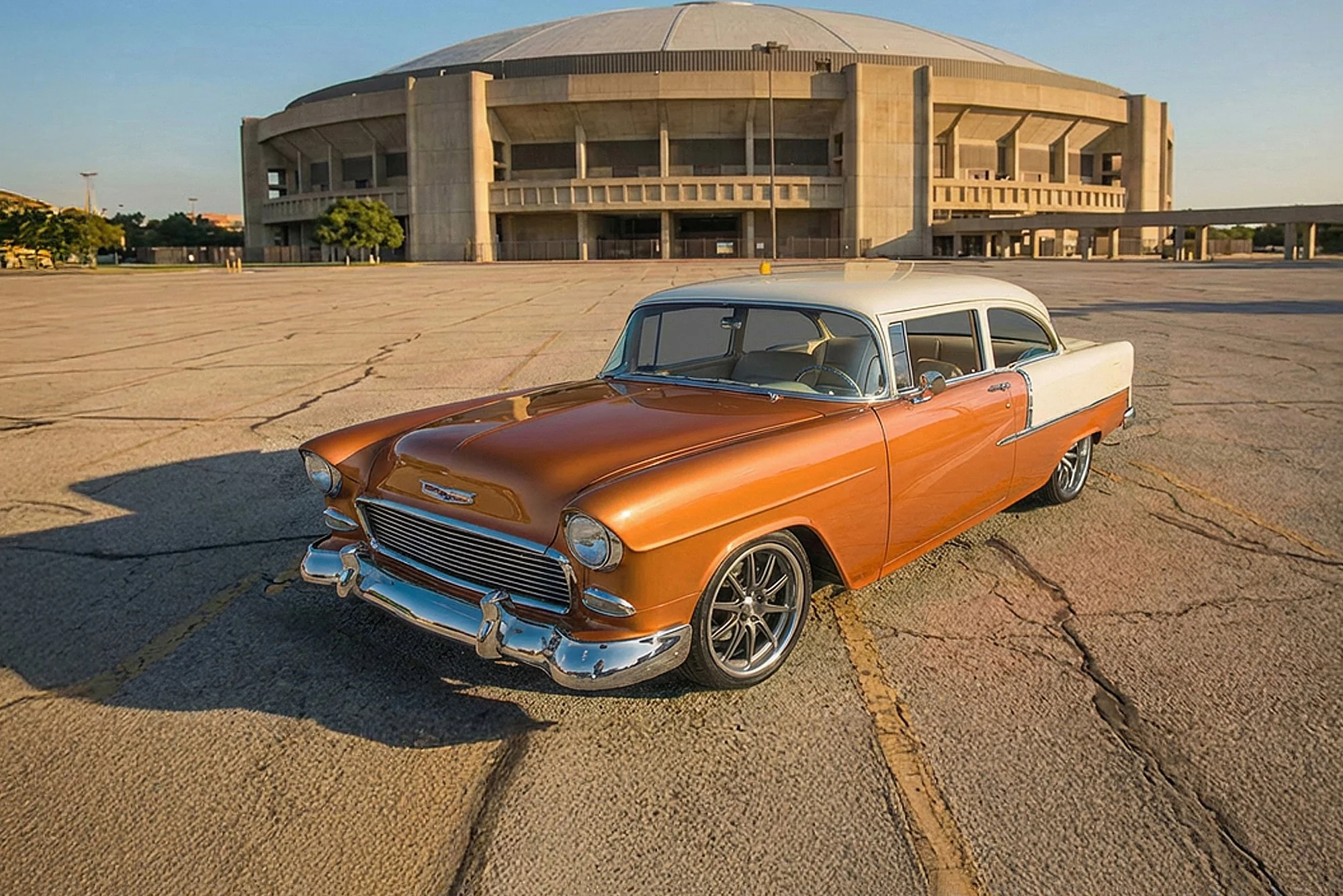 A vintage orange and white classic car parked in an empty lot with large concrete stadium in the background during sunset.