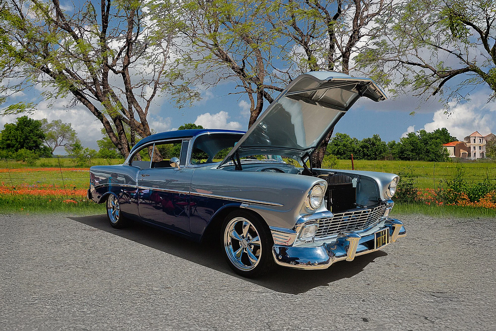 A vintage gray and blue classic car with its hood open, parked beside a large tree in a rural area with green fields, a fence, and a house in the background, under a blue sky with clouds.