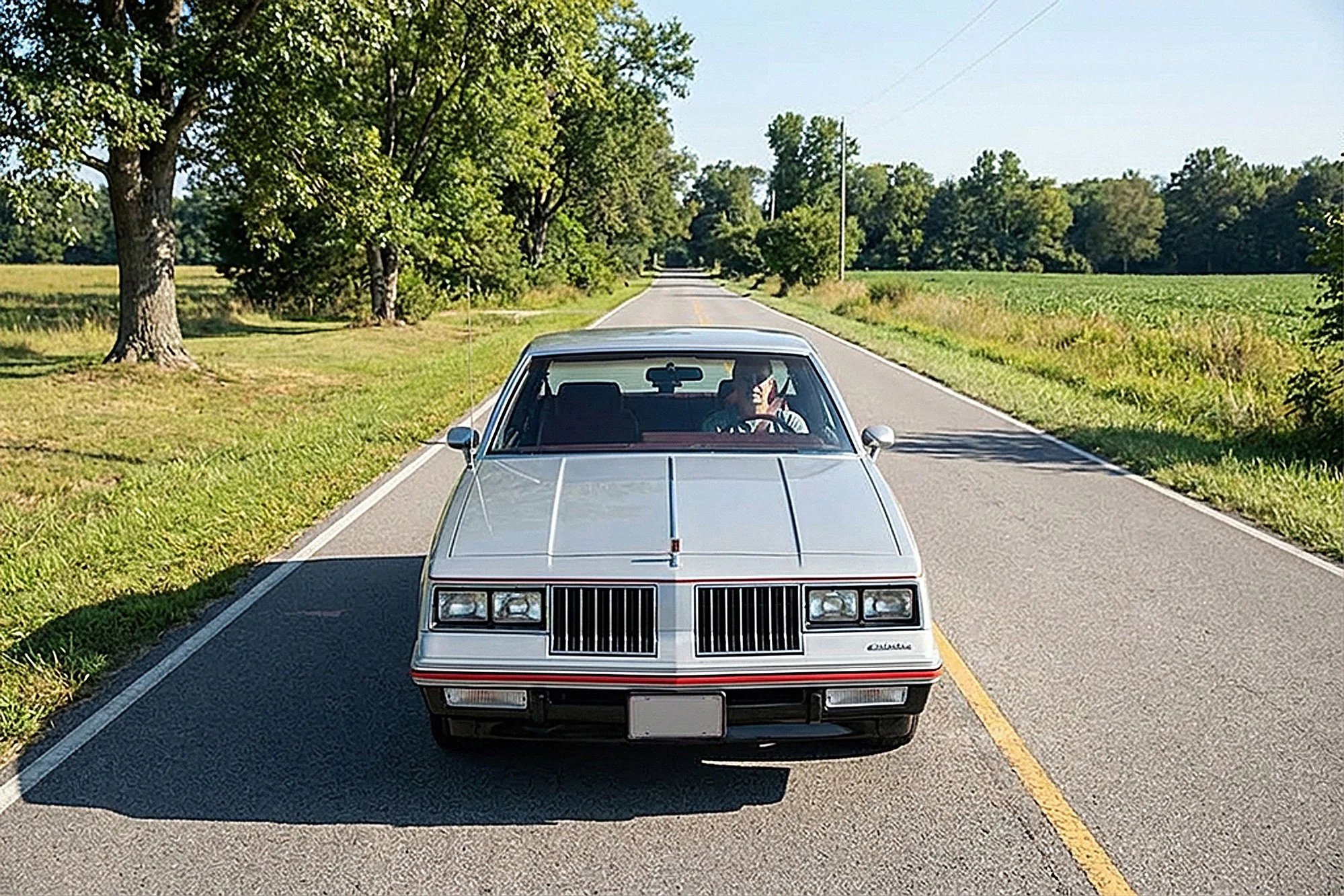 A silver DeLorean car driving on a two-lane rural road surrounded by green trees and fields, with a person visible inside the car.