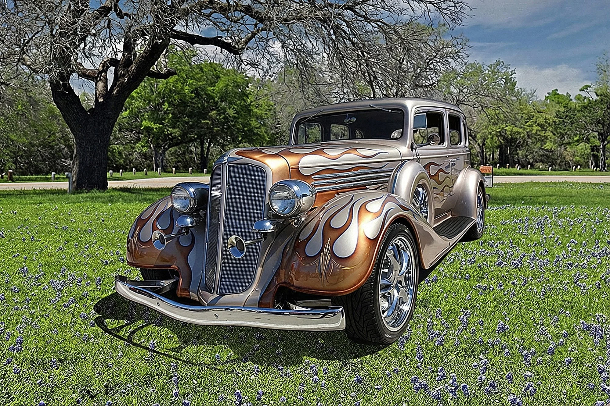 A vintage car with a flame paint job parked on a grassy field with purple flowers, under a partly cloudy sky, with trees in the background.