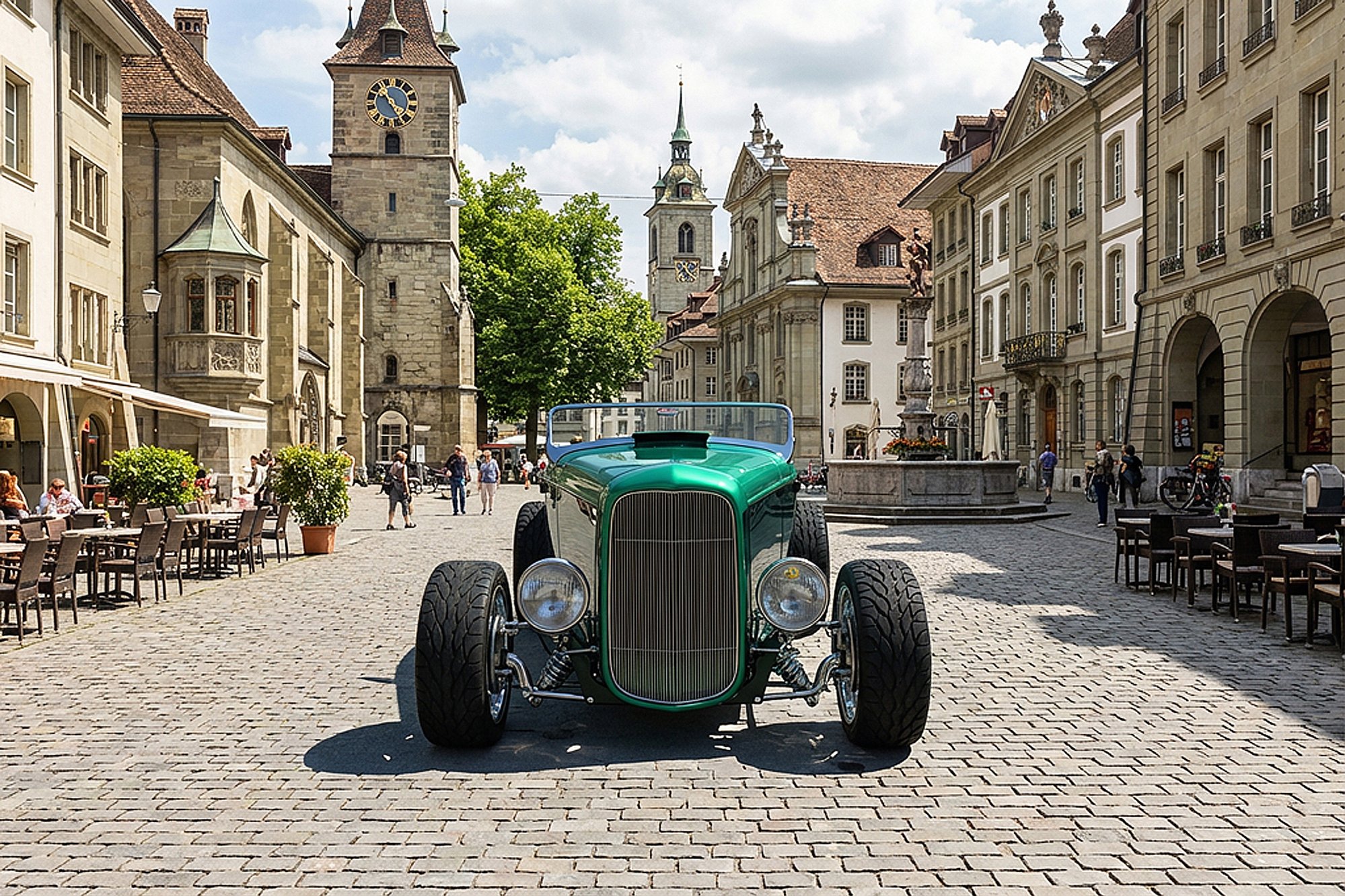 A vintage green car parked in a European city square with historic buildings, clock towers, outdoor cafes, and people walking around.