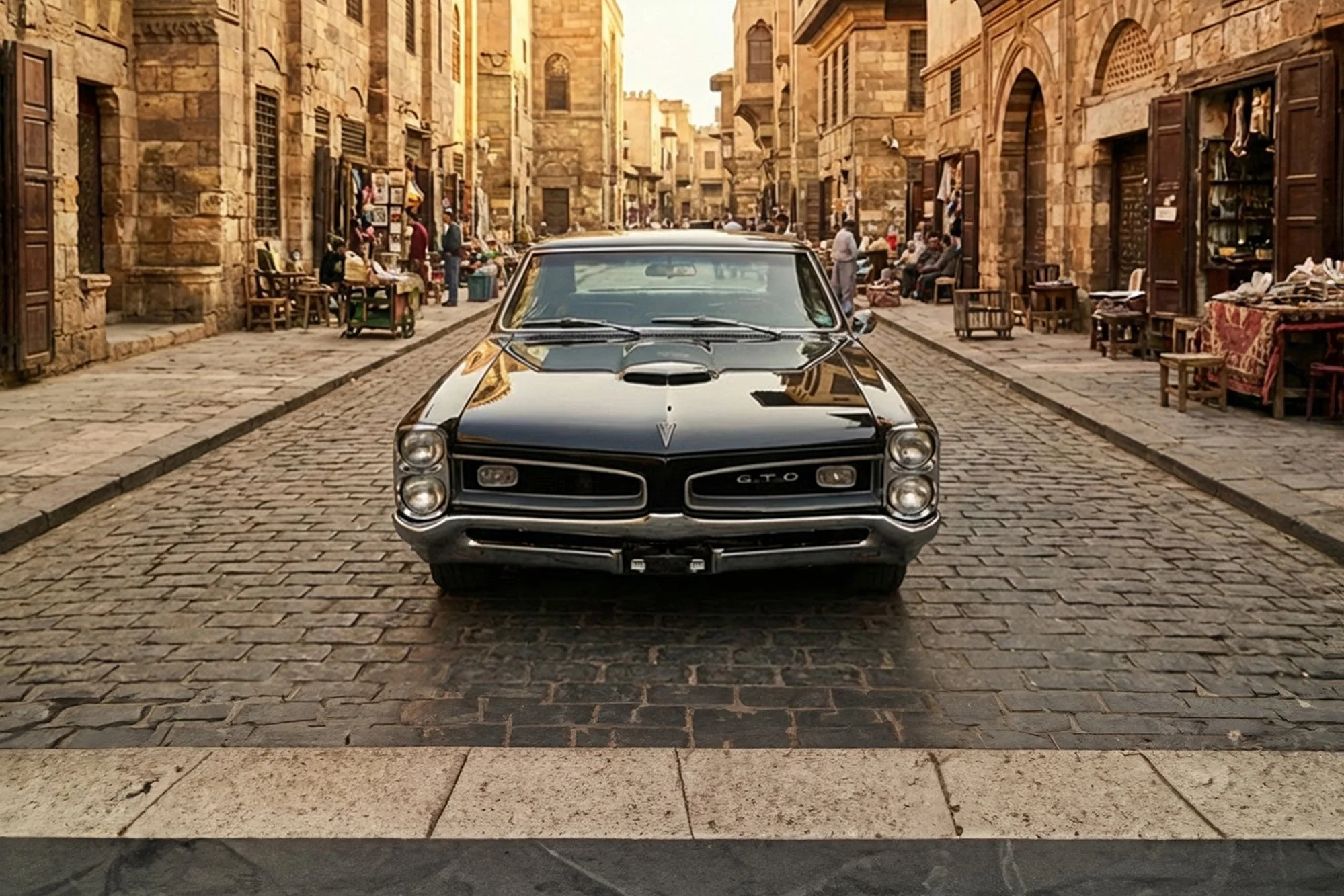 A classic black Pontiac GTO muscle car is parked on a cobblestone street in front of historic stone buildings. The street is lively with people sitting and standing, engaging in activities, with shops displaying artwork and textiles.