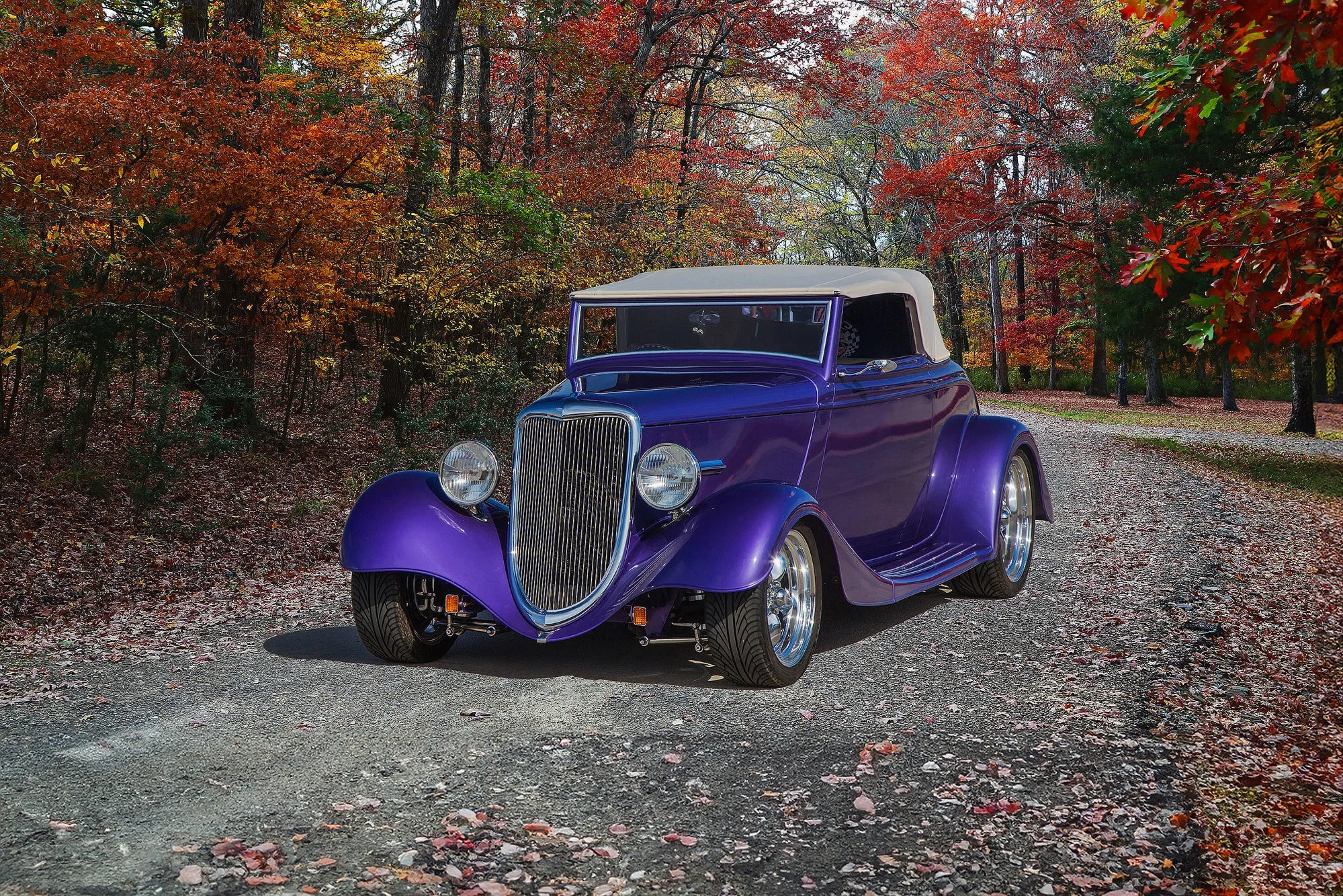 A vintage purple convertible car with a white soft top parked on a dirt road amid an autumn forest with colorful red, orange, and yellow leaves on trees and on the ground.