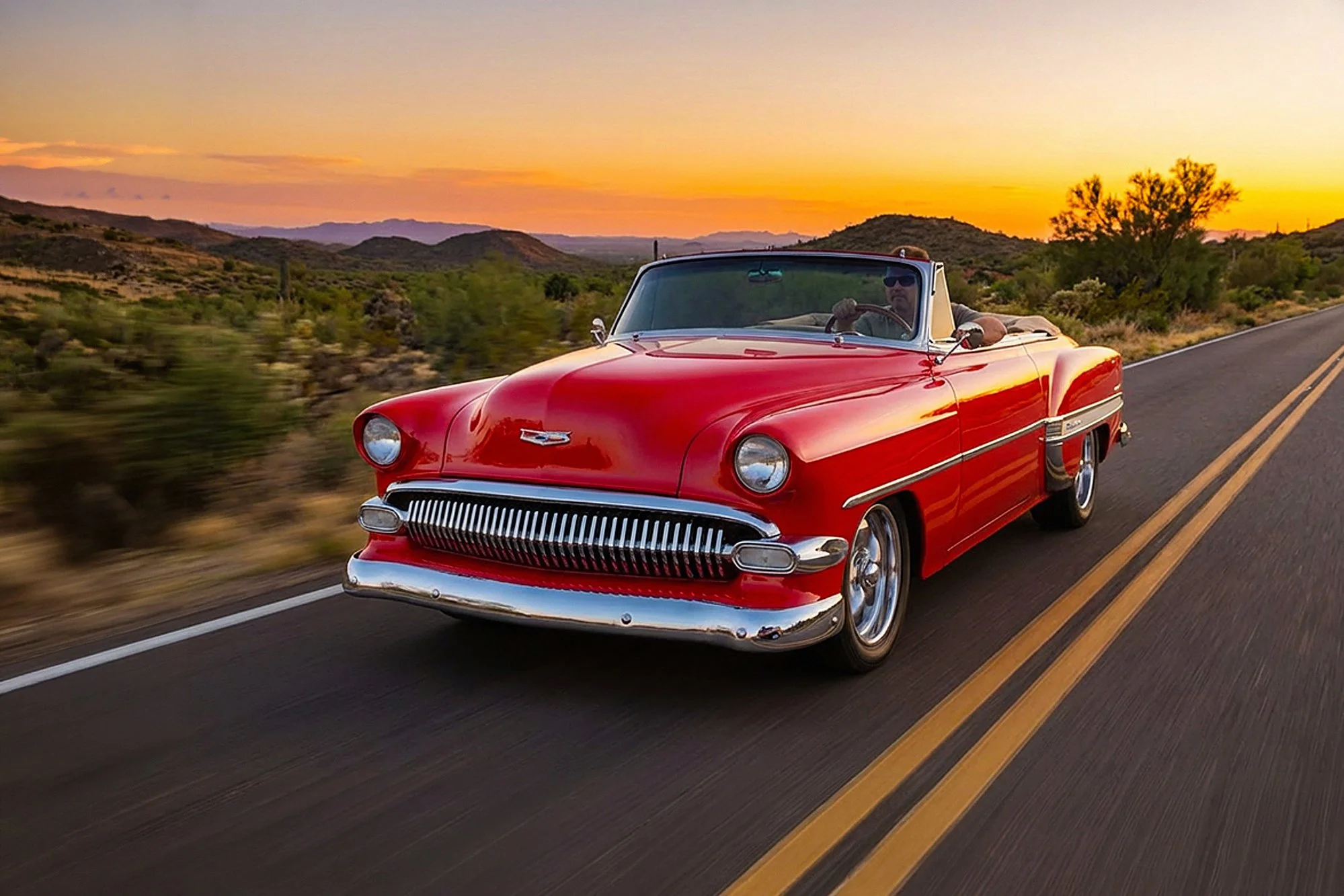 A man driving a red vintage convertible car on a two-lane road through a desert landscape at sunset.
