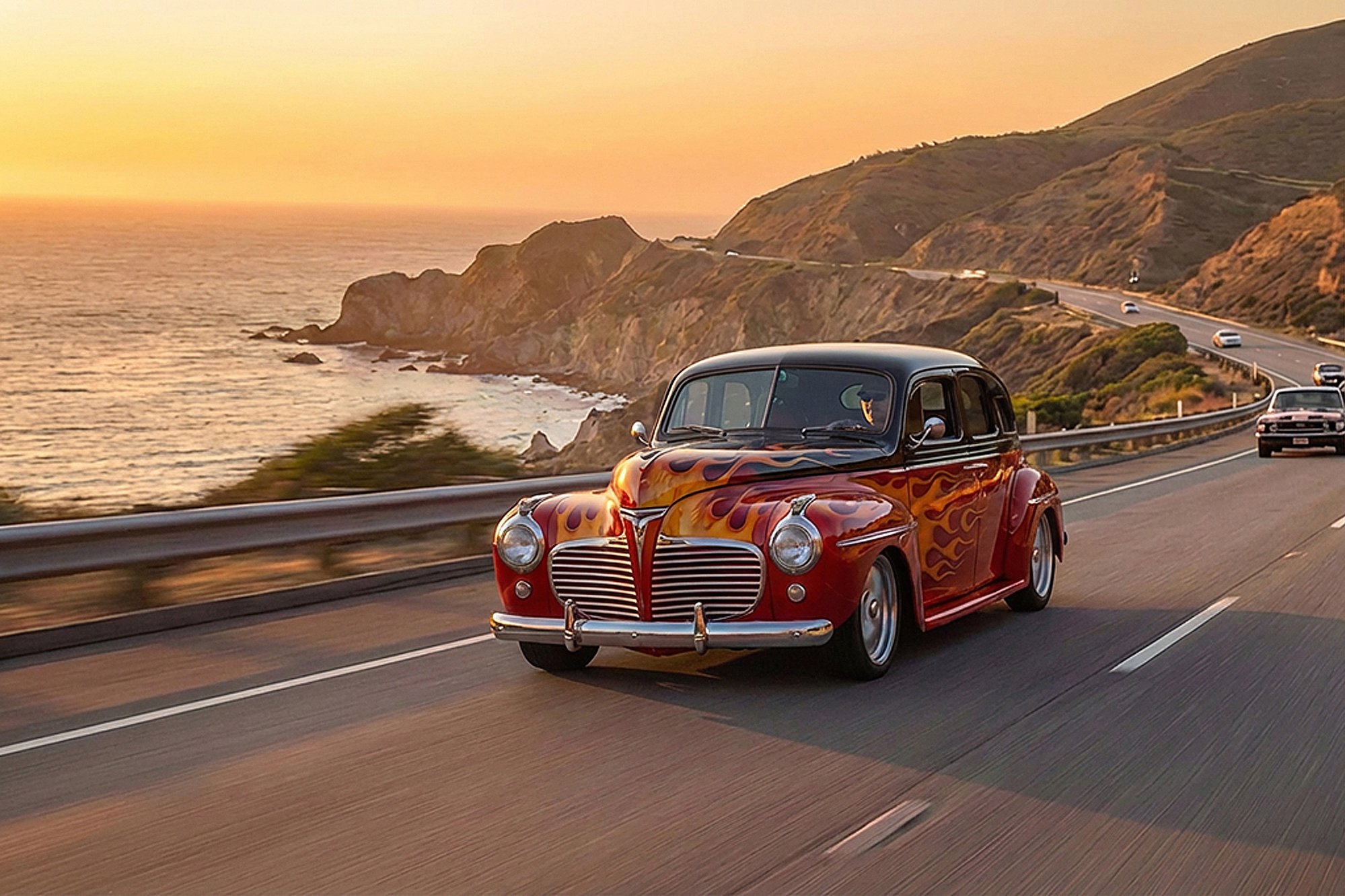 A classic vintage car with flames painted on its red body driving along a coastal highway during sunset.