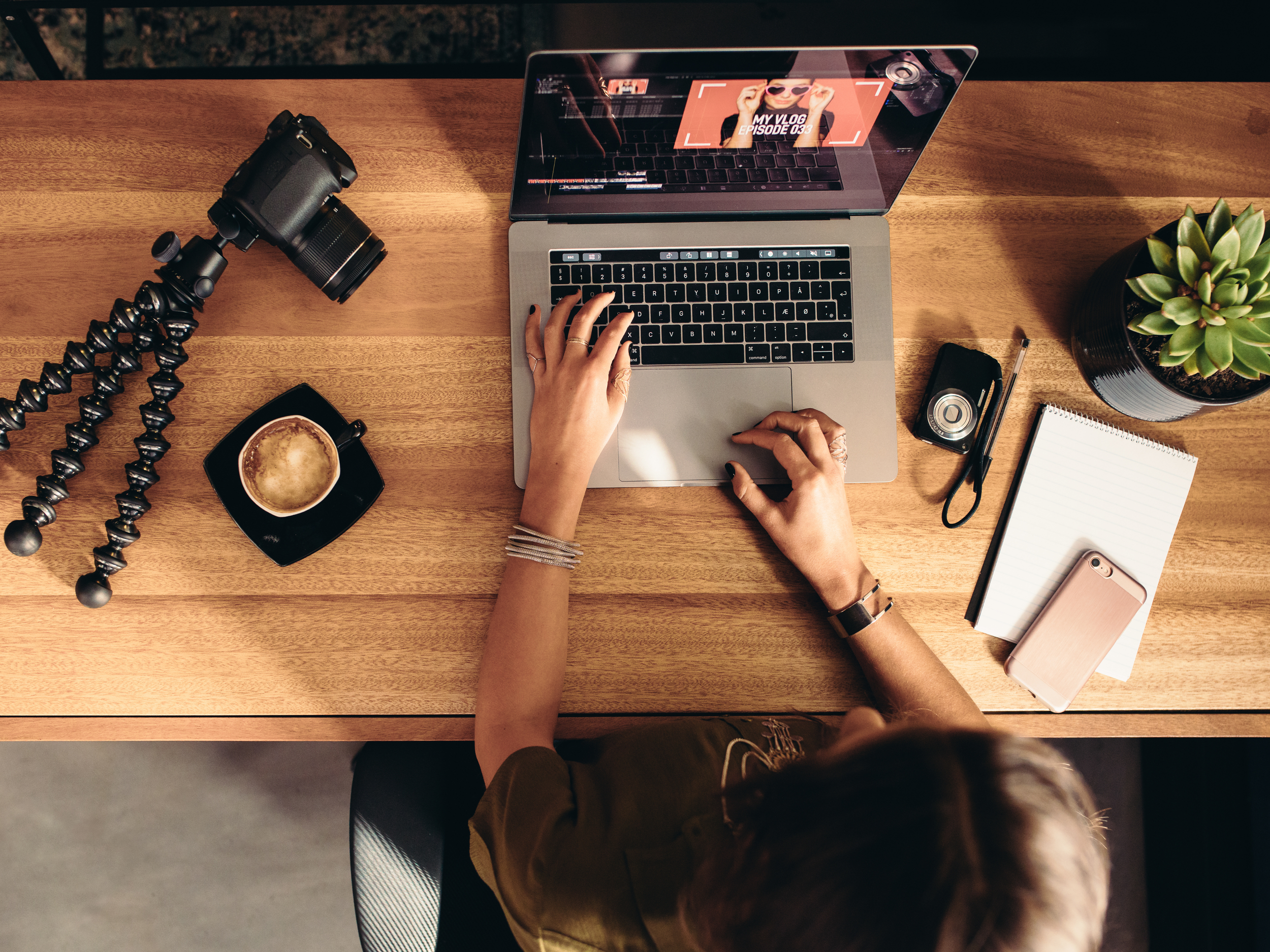Ein Holzschreibtisch mit einer Person, die an einem Laptop arbeitet, einem Kamera-Stativ, einer Tasse Kaffee, einer Kamera, einem Notizbuch, einem Smartphone, einem Zollstock, einem Notizblock mit Stift, einer Pflanze und einer Kamera auf dem Tisch.