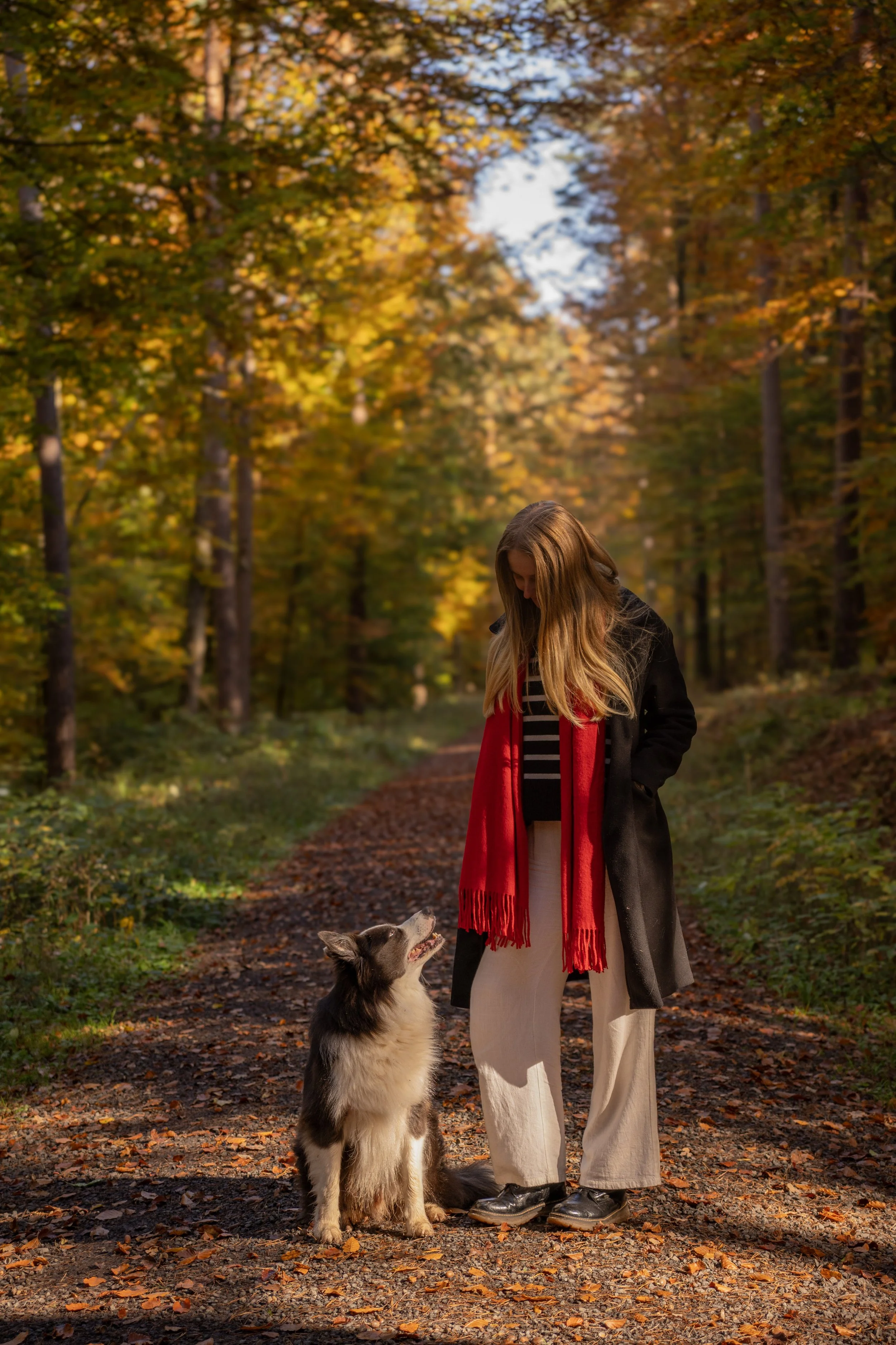 Frau mit langem Haar, dunkler Jacke und roter Schal, steht in einem herbstlichen Wald mit Hund, auf einem von Blättern bedeckten Weg.