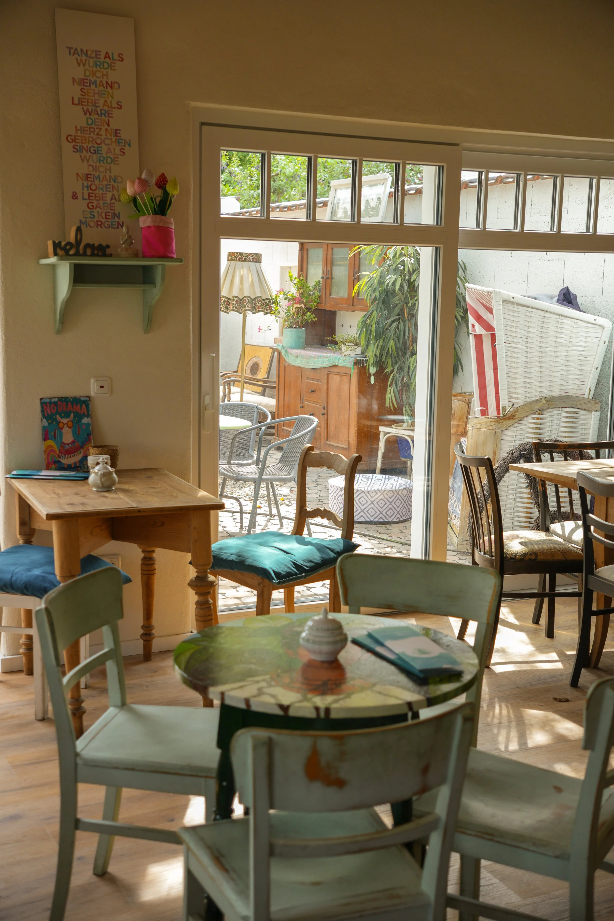 Ein Blick in ein gemütliches Café mit einem kleinen Tisch, mehreren Stühlen und einem großen Fenster, das auf einen Garten im Freien blickt, mit Pflanzen, Gartenmöbeln und einem Sonnenschirm.