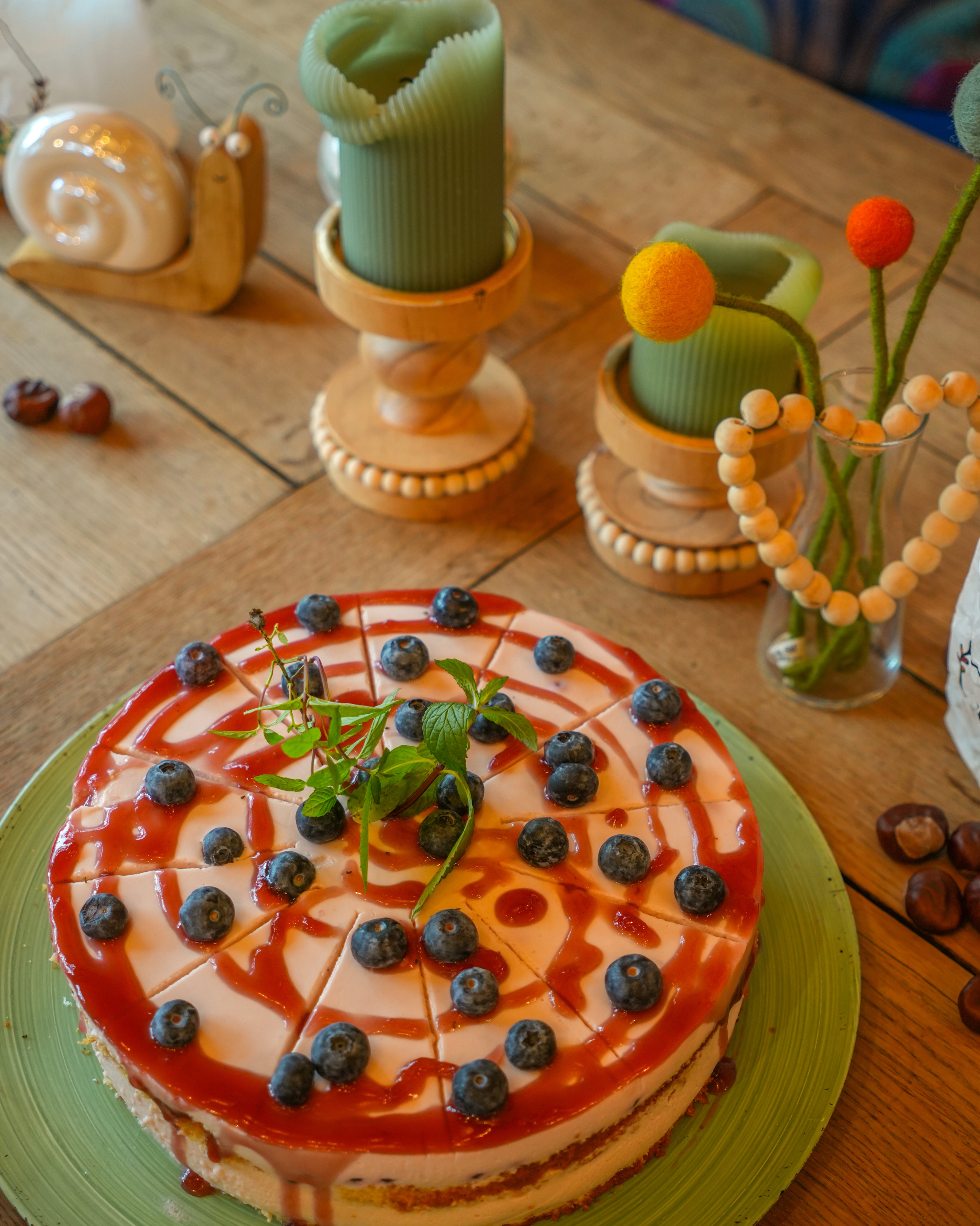 Ein Kuchen mit Blaubeeren und Minze auf einem grünen Teller, umgeben von Dekorationen wie Kerzen, Schneckenfiguren, einem Herz aus Holzperlen und getrockneten Feigen auf einem Holztisch.