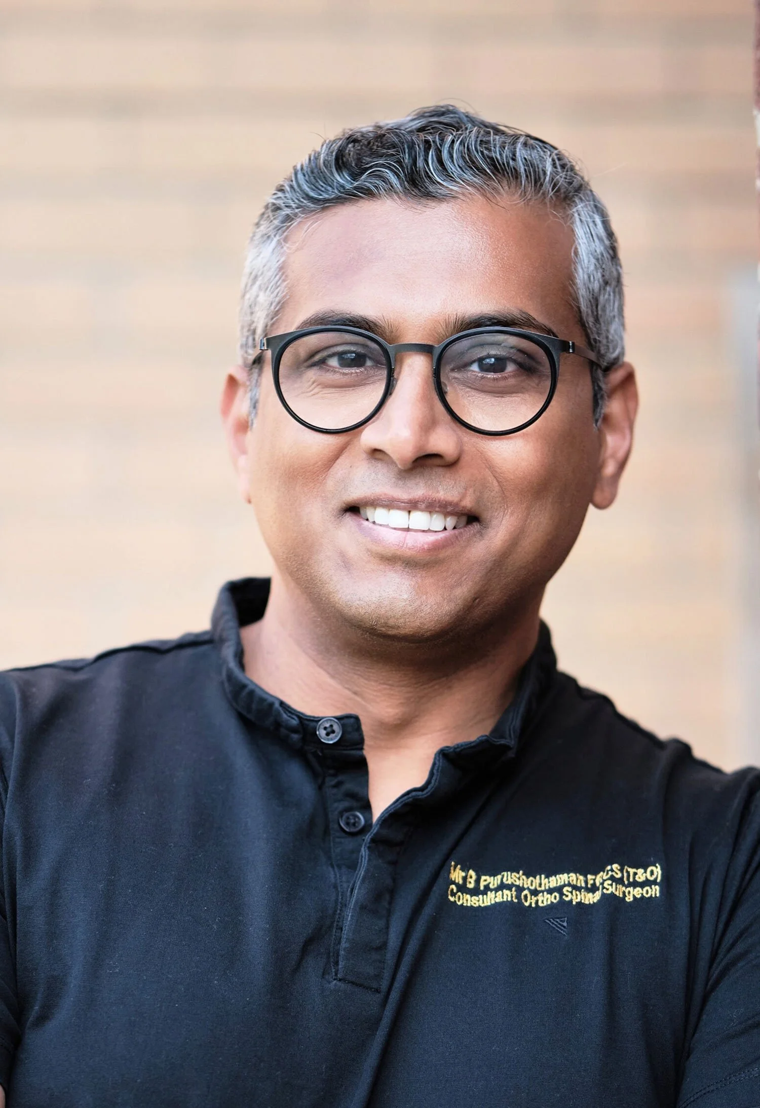 A smiling man wearing glasses and a black shirt with yellow embroidered text stands against a beige brick wall.