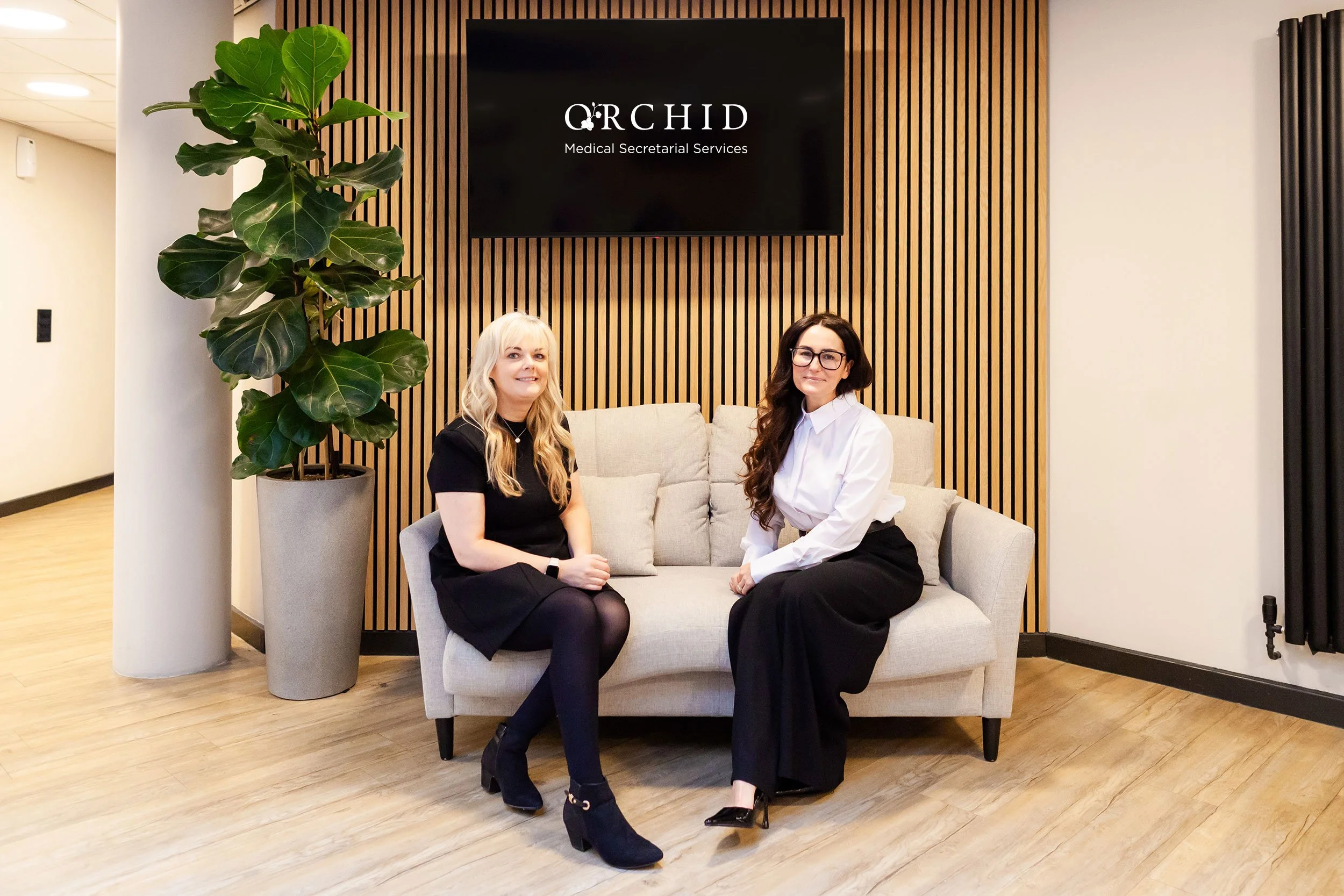 Two women sitting on a beige couch in a modern office lobby, with a large green plant on the left, a wall-mounted TV displaying 'Orchid Medical Secretarial Services' behind them, and wooden paneling on the wall.
