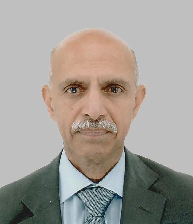 A middle-aged man with a mustache, wearing a dark suit, light blue shirt, and a patterned tie, against a plain gray background.