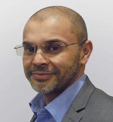 Close-up of a man with glasses and a beard, wearing a suit and blue shirt, smiling subtly.