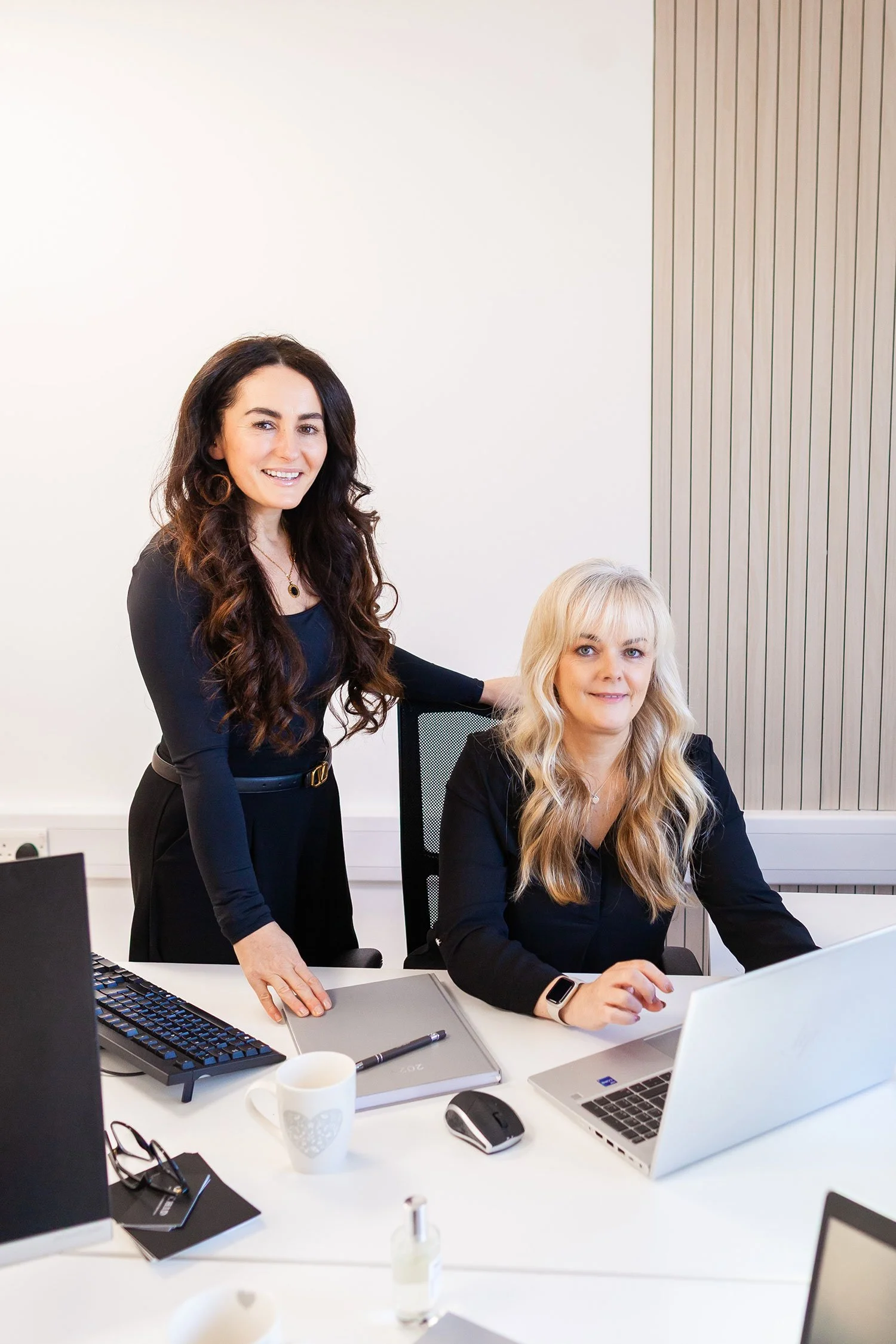 Two women working in an office. One woman with dark, long, curly hair is standing and smiling, while the other woman with blonde, wavy hair is sitting at a desk with a laptop. The desk has a keyboard, mouse, glasses, a notebook, a mug, and a hand sanitizer bottle.