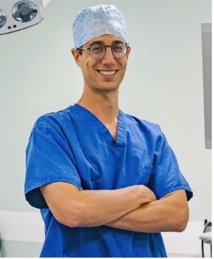 A male surgeon in blue scrubs, a surgical cap, and glasses, smiling with his arms crossed in a medical setting.