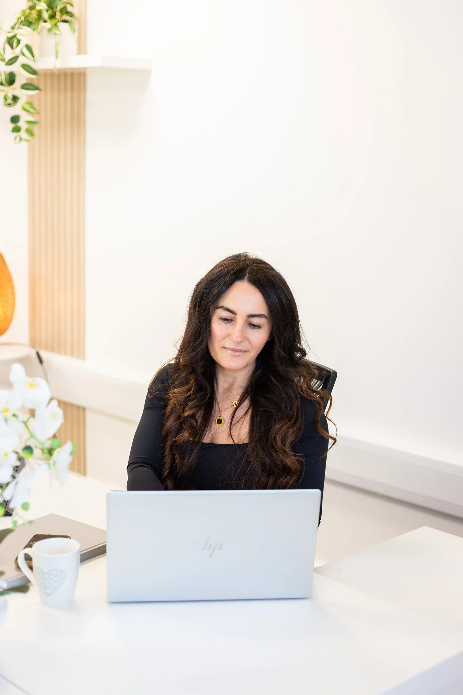 A woman with long dark hair sits at a white desk working on a silver laptop in a bright, modern office, with a white mug and a potted plant nearby.