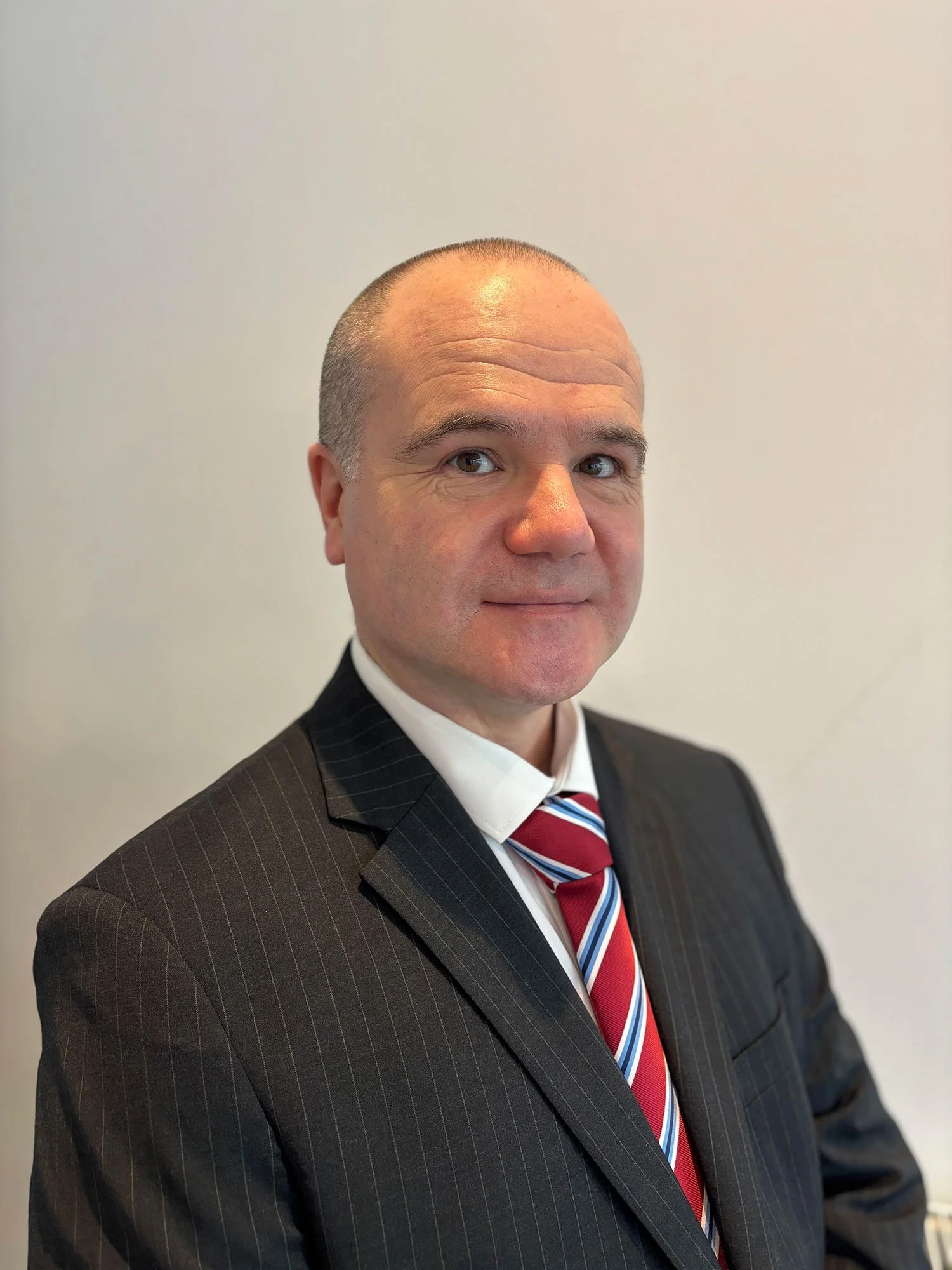 A middle-aged man wearing a black pinstripe suit, white shirt, and a red, white, and blue striped tie, standing against a plain light-colored wall.