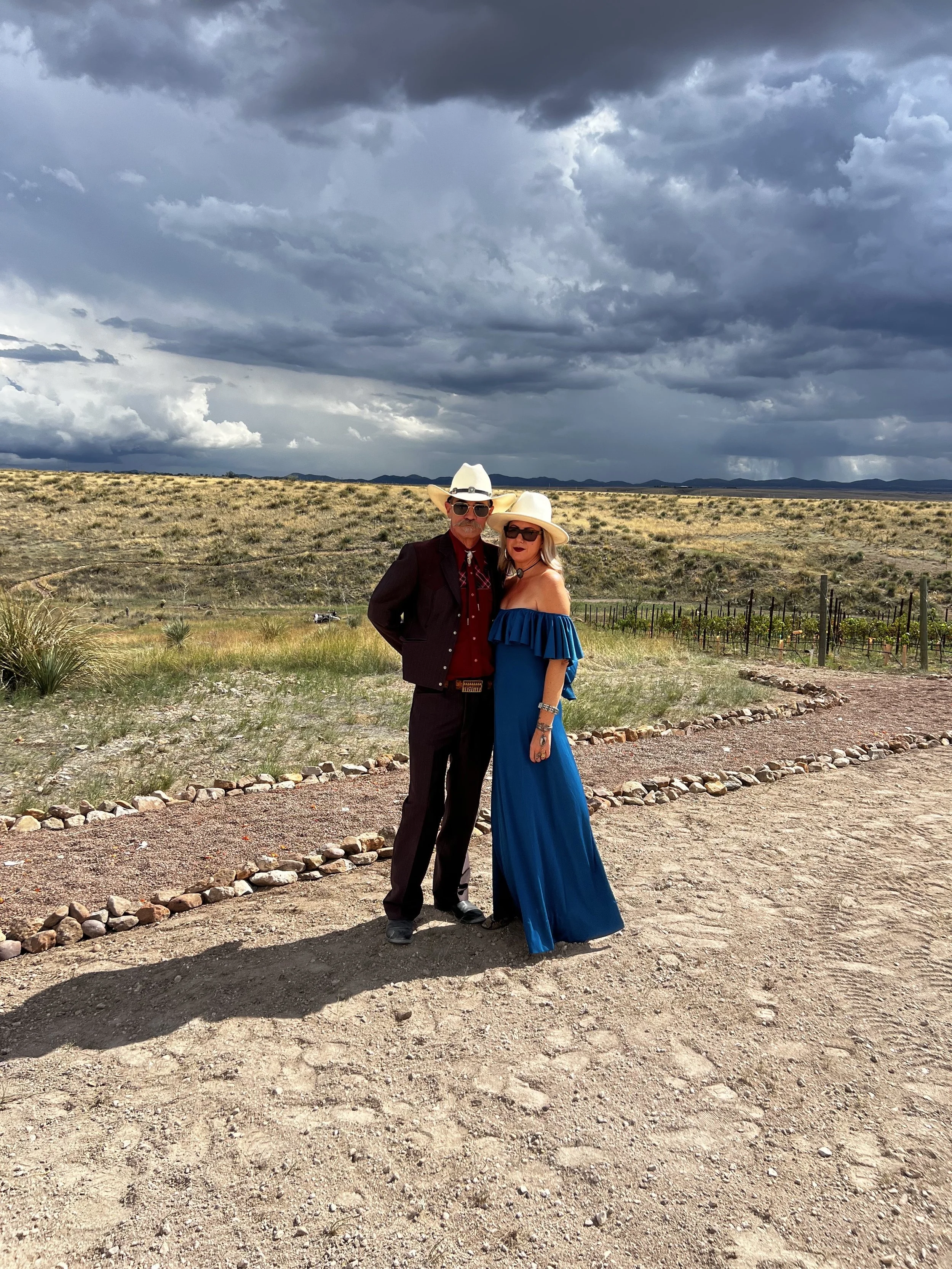 A man and woman dressed in Western-style clothing standing on a dirt path with a cloudy sky overhead and a desert landscape in the background.