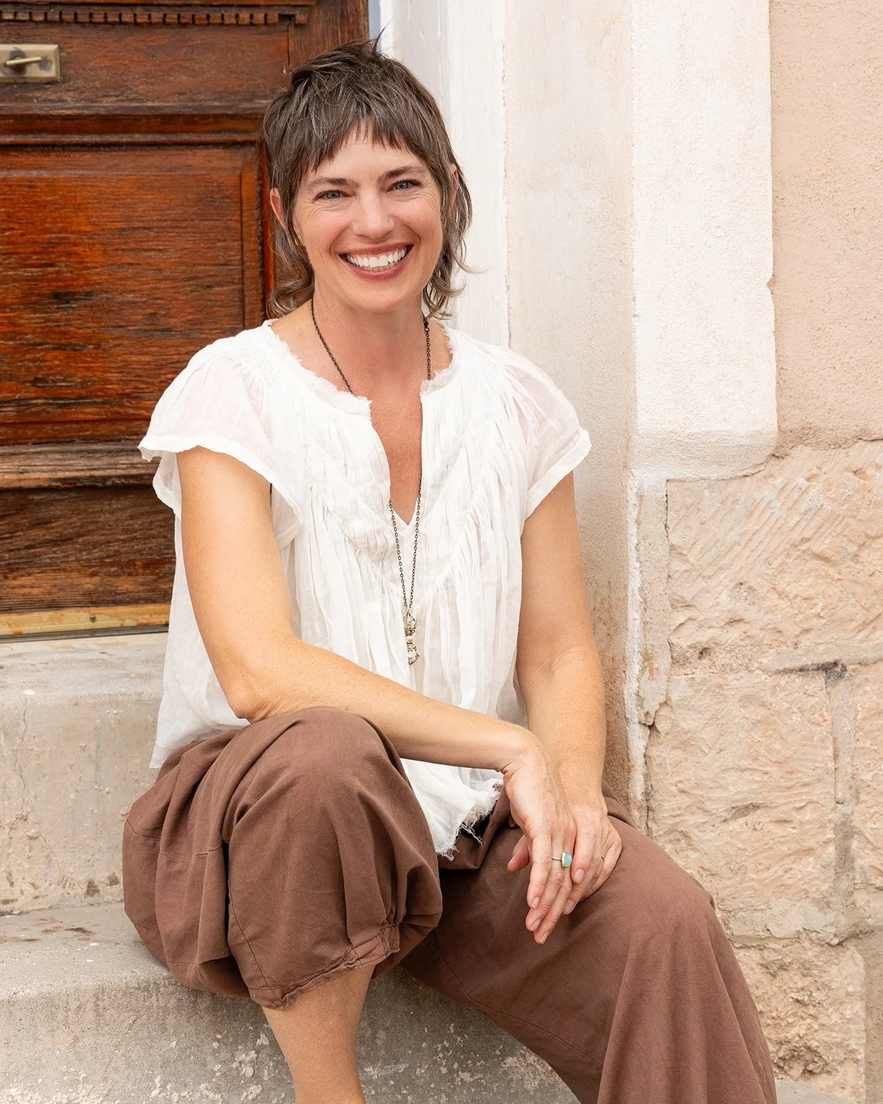 A woman sitting on steps outside, smiling, wearing a white short-sleeved top and brown pants.