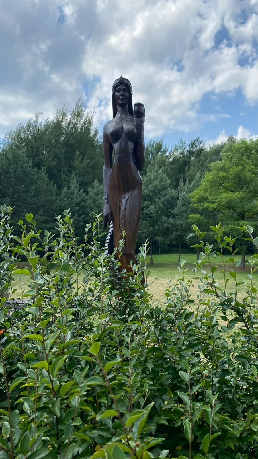 A tall wooden sculpture of a woman holding a cup, situated outdoors among green bushes and trees, with a partly cloudy sky in the background.