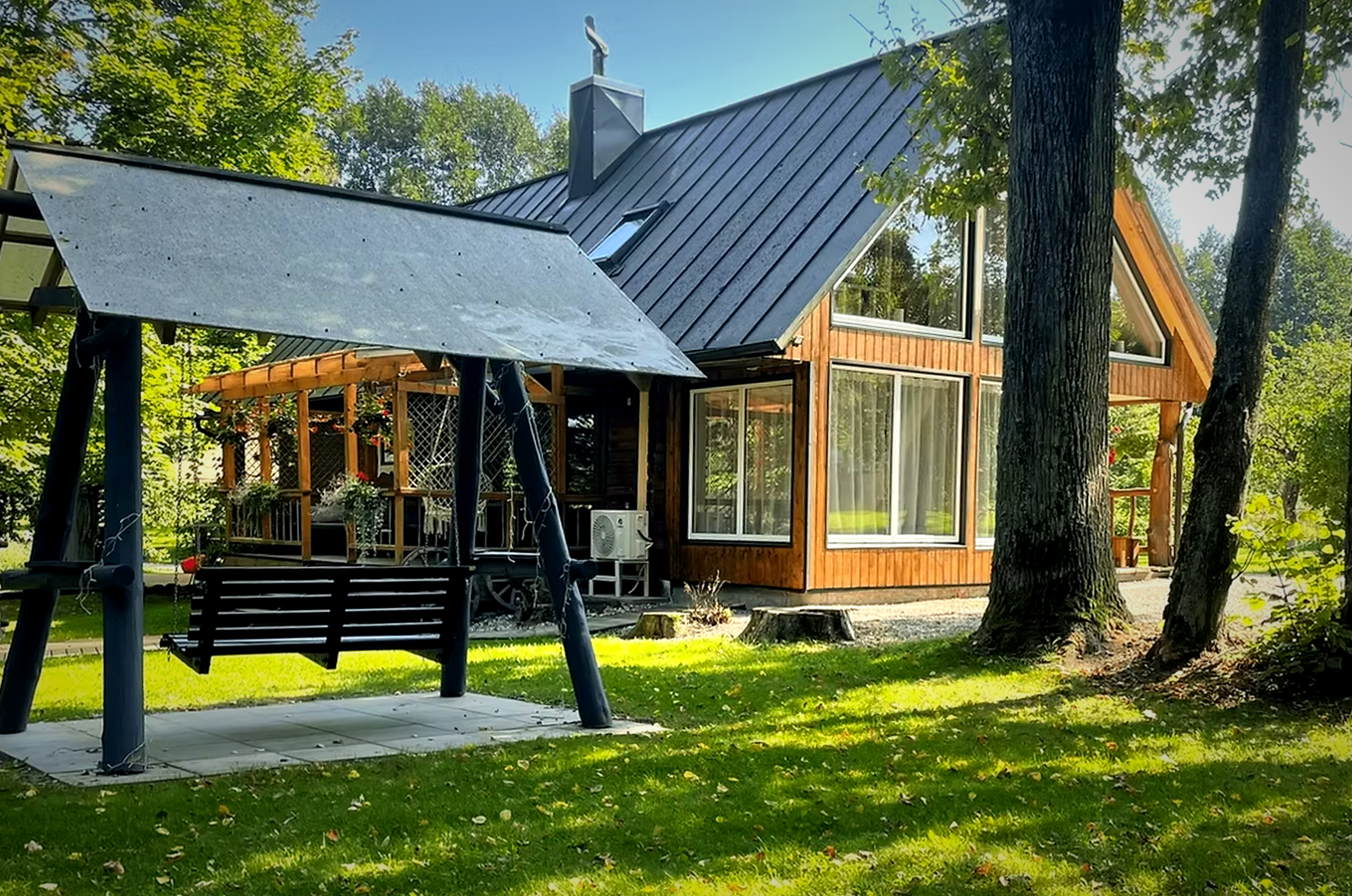 A house with large windows and a wooden exterior, surrounded by trees and greenery, with an outdoor patio area and swing set in the yard.