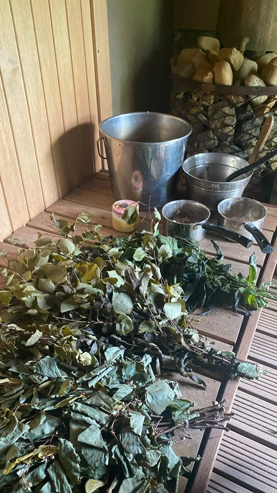 A wooden table with freshly cut green leaves and sauna whisks, stainless steel buckets, small pots, and a container of honey, set in a sauna room with wooden walls and an arrangement of sauna rocks in the stove.