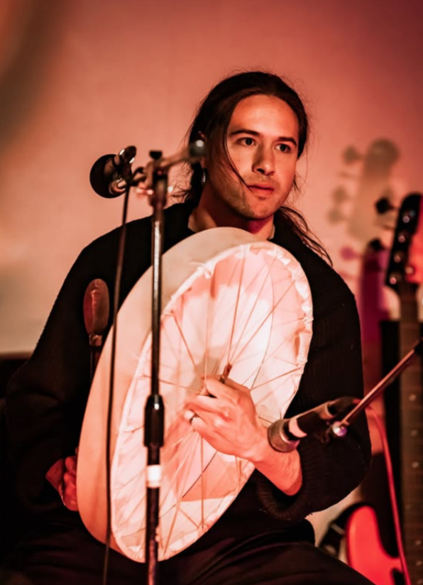 Musician sitting with a percussion instrument, holding a drum and a mallet, in front of microphones and musical equipment.