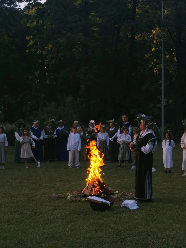 People in traditional Lithuanian clothing gathered outdoors around a campfire in a grassy area with trees in the background, possibly for a Rasos midsummer ceremony