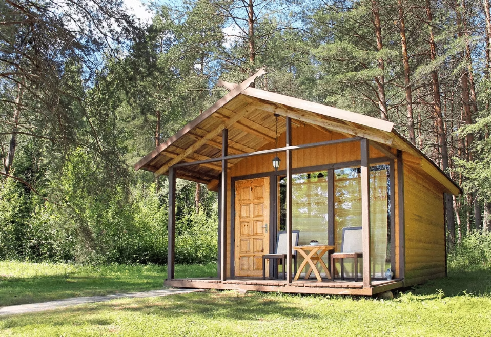 Small wooden cabin in the woods with a porch, two chairs, and a small table, surrounded by greenery and trees.