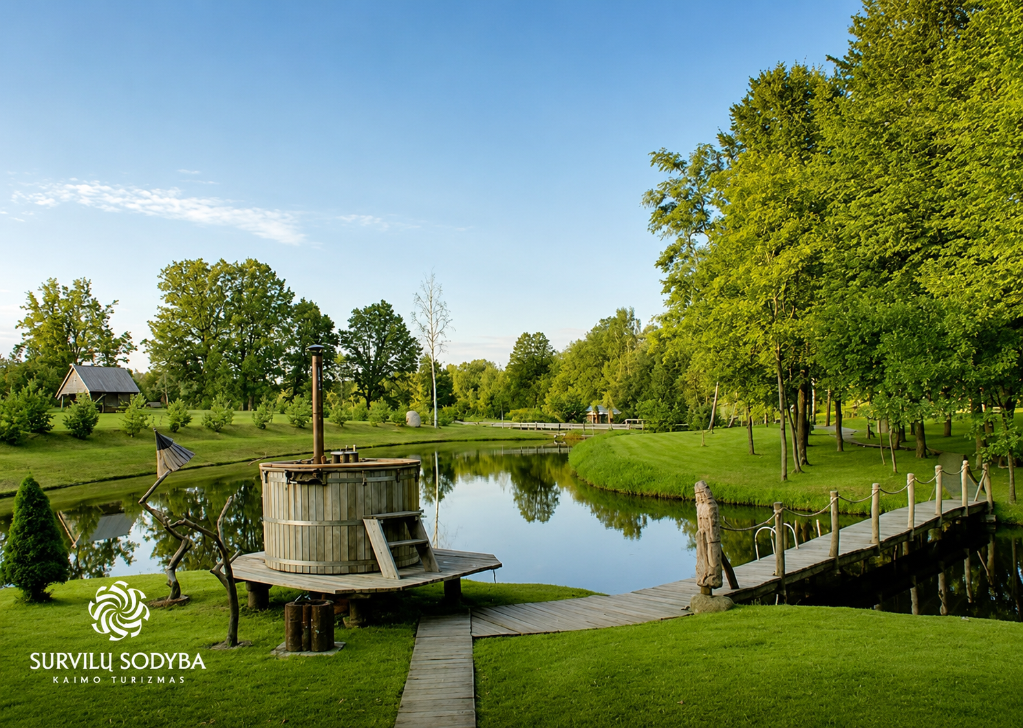A wooden bridge over a narrow lake in a homestead with lush green grass and tall trees, with a barrel-shaped wooden hot tub and a unique metal sculpture nearby, under a clear blue sky.