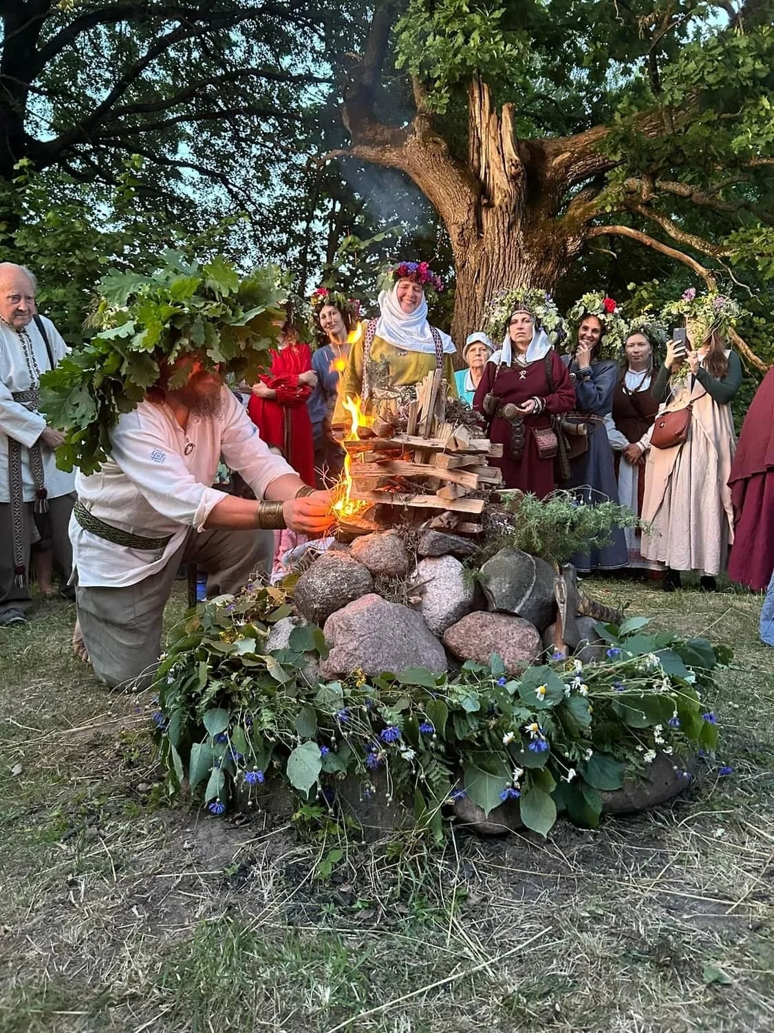 People dressed in traditional clothing gathered outdoors around a decorated stone and wood fire, with some women wearing flower crowns, participating in a cultural or ceremonial event under a large tree.