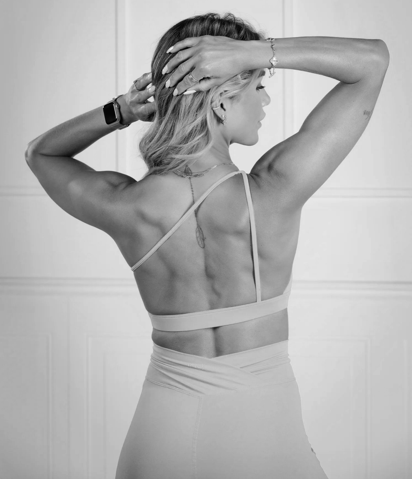 A woman with fit physique in workout attire, lifting her hair, standing sideways, wearing jewelry and a smartwatch, in front of a plain background.