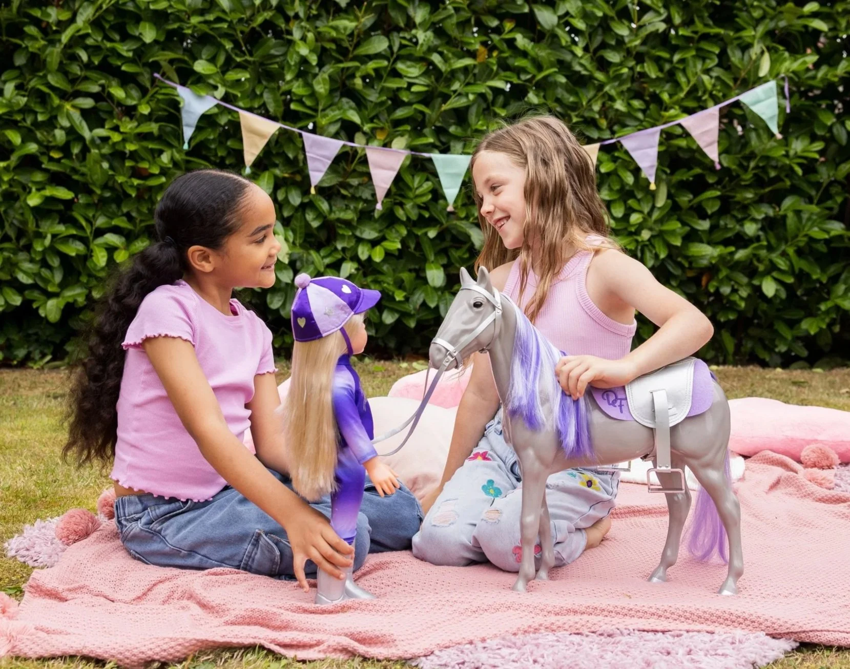 Two girls playing outside, smiling at each other. Playing with their 18 inch DesignAfriend doll dressed in a riding outfit and a grey plastic horse.