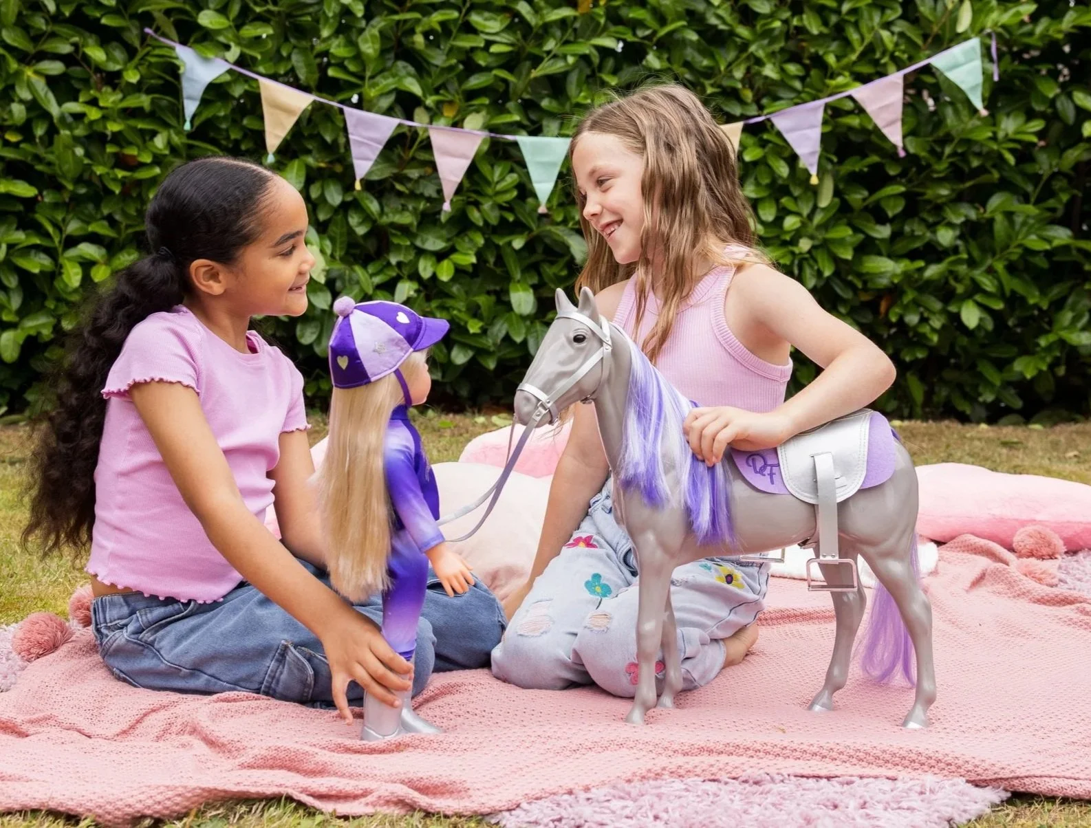 Two young girls outside in the garden smiling to each other,  playing with their DesignAfriend doll and horse playset.
