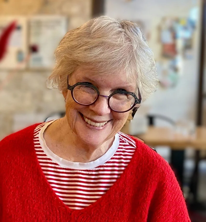 A smiling older woman with short, gray hair, wearing glasses and a red sweater over a white and red striped shirt sits in a cozy indoor setting.