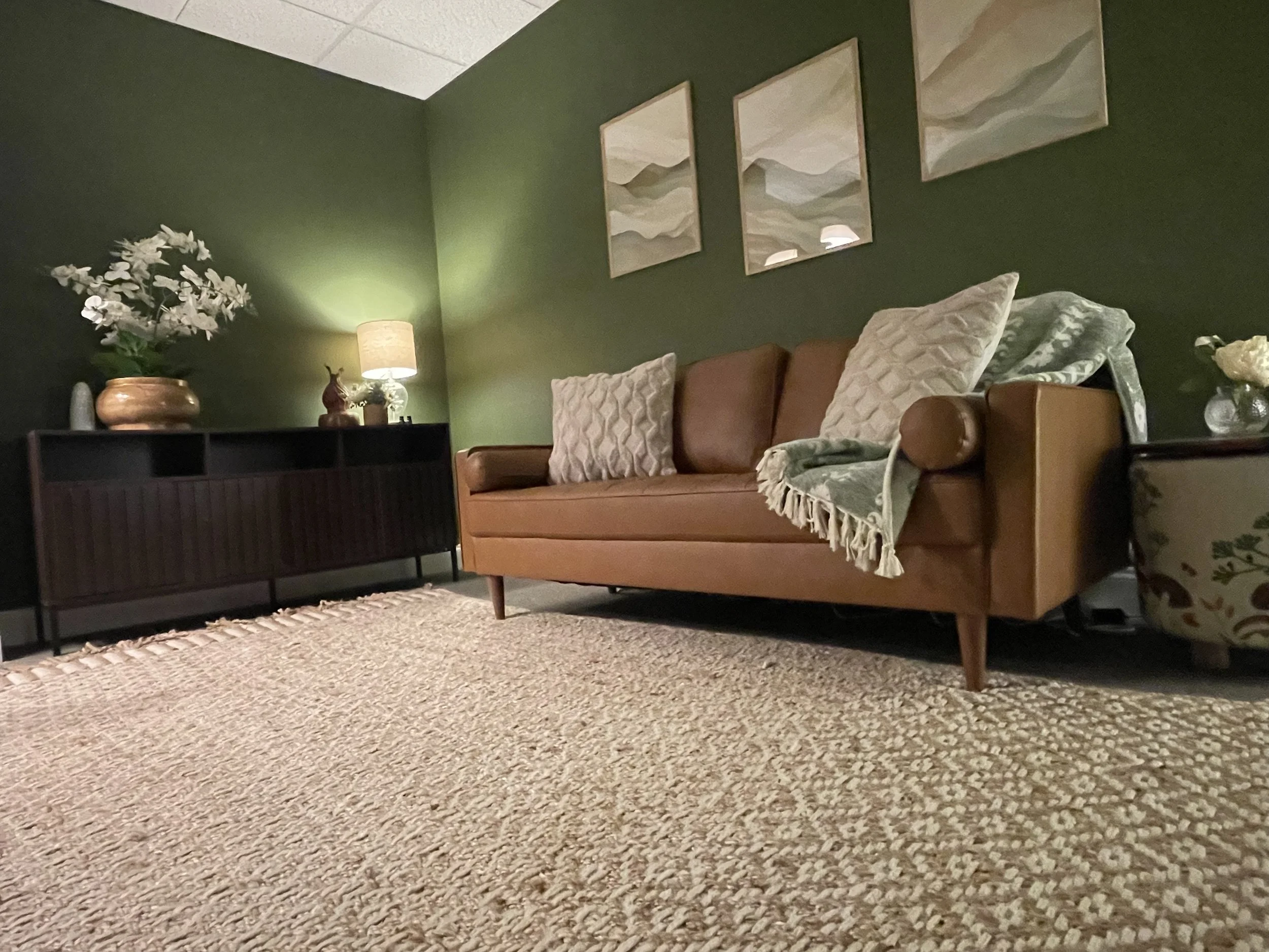Living room with a green wall, a tan sofa with decorative pillows and a throw blanket, a black sideboard with a vase of white flowers and a lamp, a textured beige carpet, and three abstract landscape paintings above the sofa.