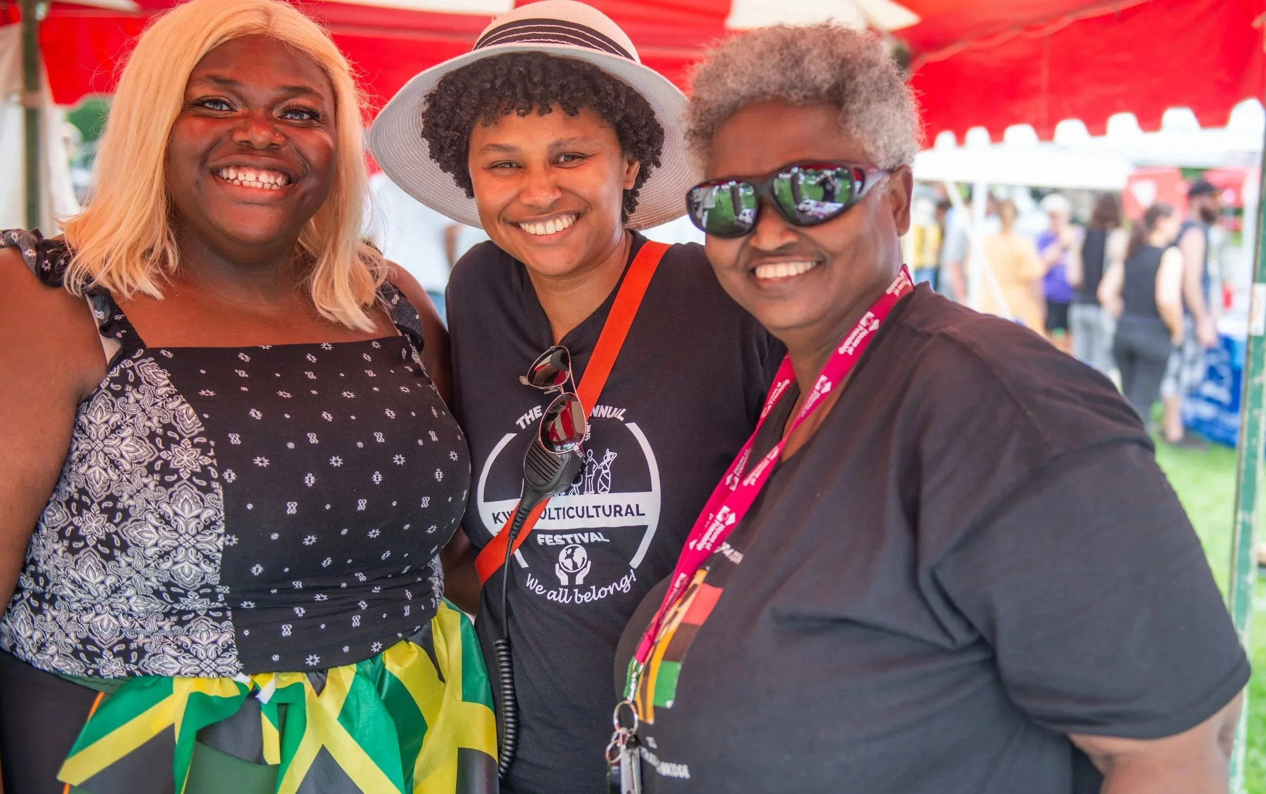 Three smiling women posing together at a cultural festival under a red tent, with a crowd in the background.