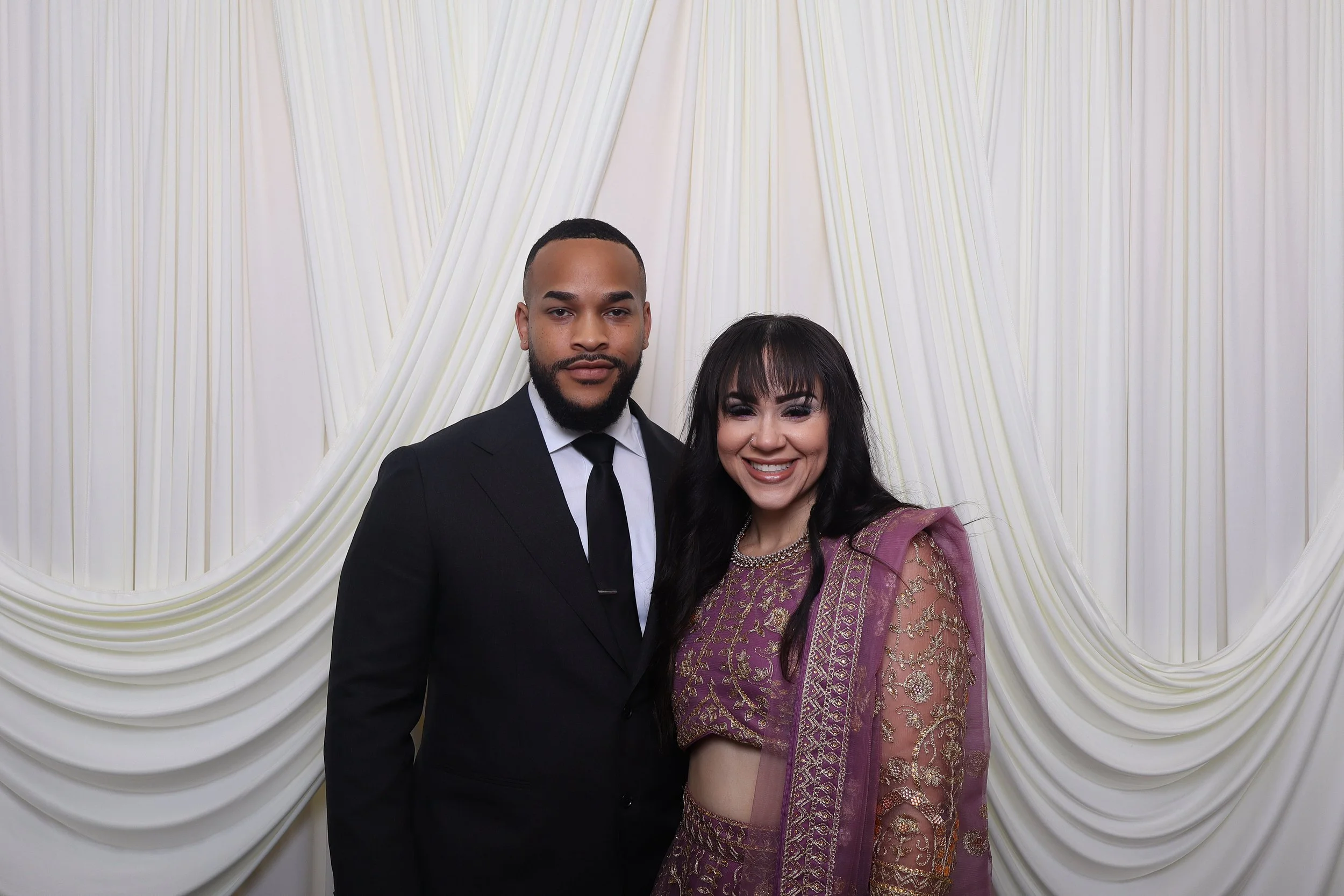 A smiling man in a black suit and tie standing next to a woman in a purple traditional dress with gold embroidery, in front of a white fabric backdrop with draped folds.
