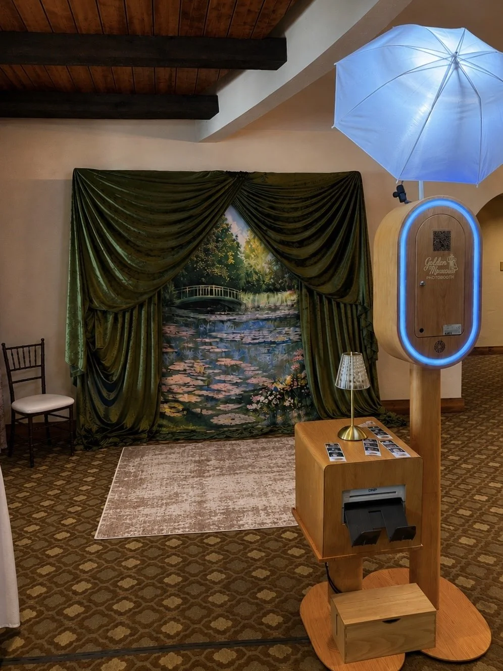 Photo of a vintage photo booth with a painted backdrops of a pond with lily pads, water lilies, and a bridge surrounded by trees, framed by dark green velvet curtains. There is a wooden table with photos, a lamp, a blue umbrella, and a QR code.
