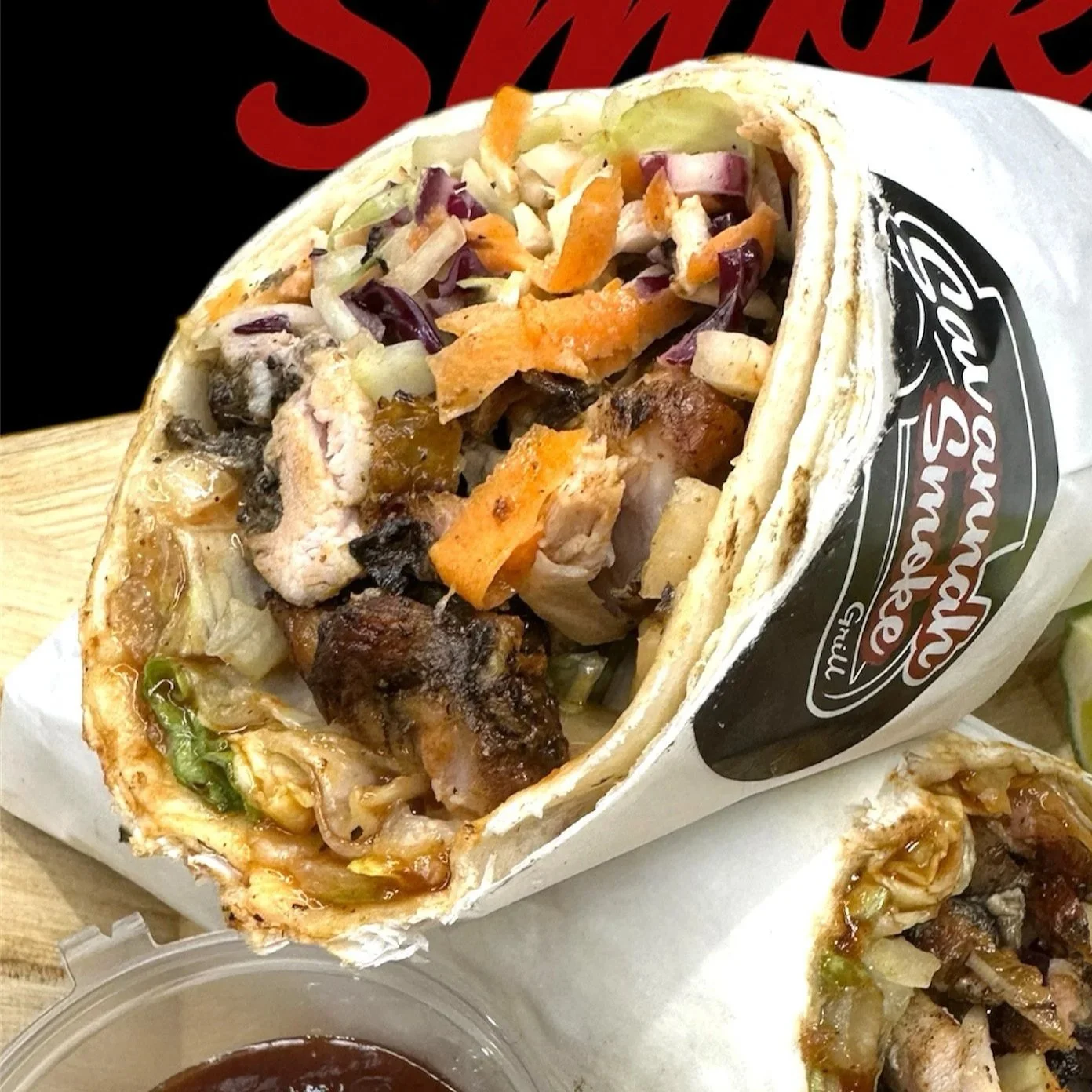 Half of a burrito wrapped in paper, filled with vegetables, grilled chicken, and meat, on a wooden tray with a small container of sauce.