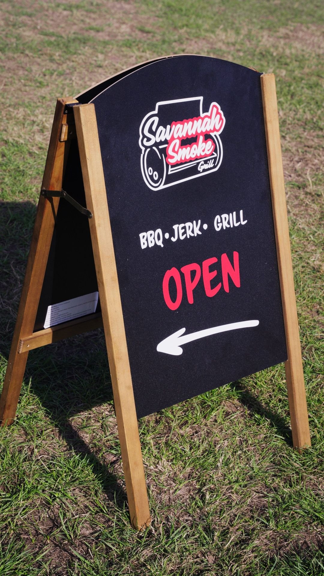 A chalkboard sign outside on grass advertising Savannah Smoke Grill, offering BBQ, jerk, and grilled food, with an arrow pointing left and the word 'Open' in red.